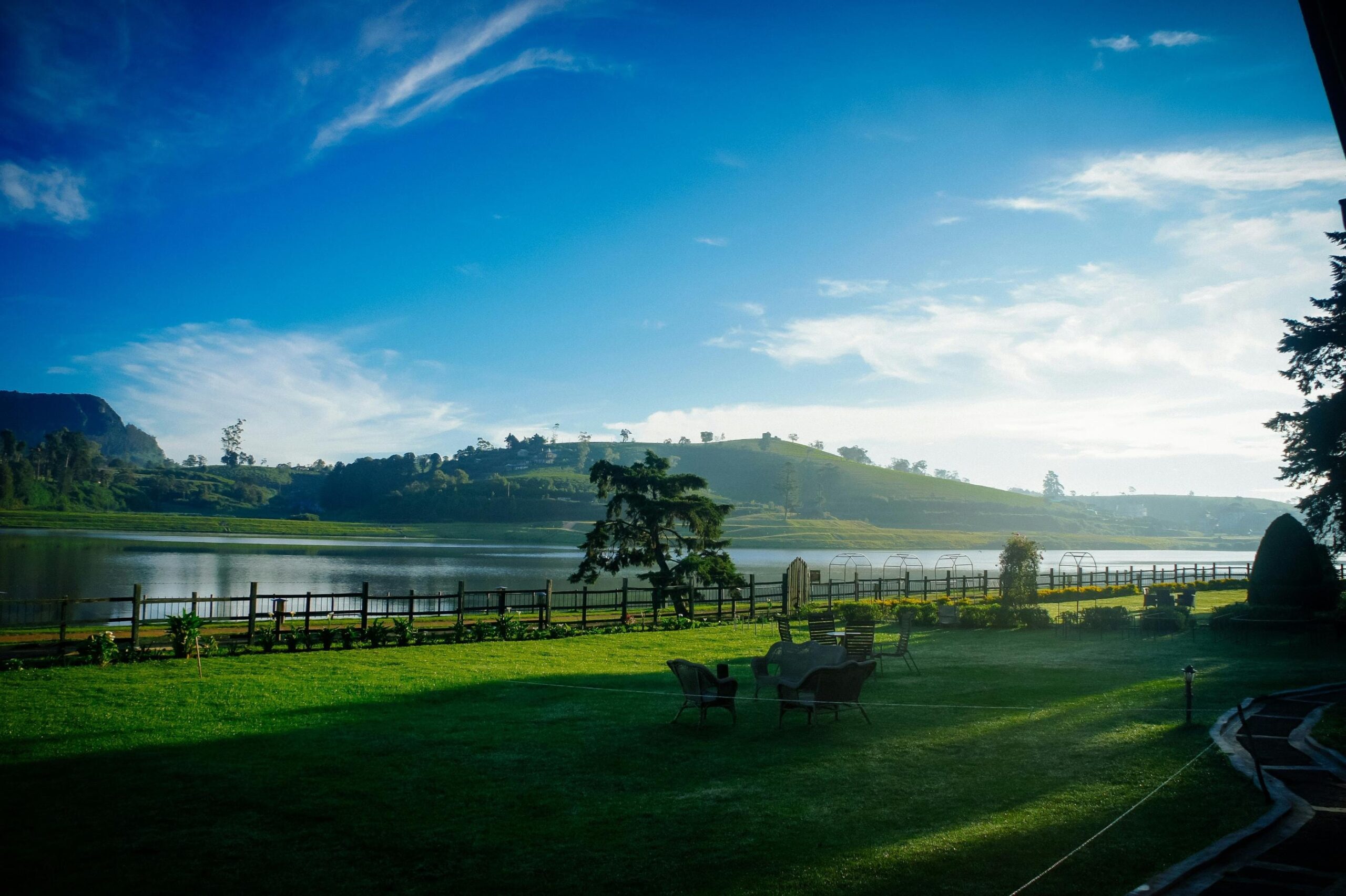 A lush green lawn dotted with outdoor seating stretches towards a calm lake, separated by a rustic wooden fence. In the background, rolling hills covered in vegetation rise beneath a vibrant blue sky filled with sweeping, wispy clouds.