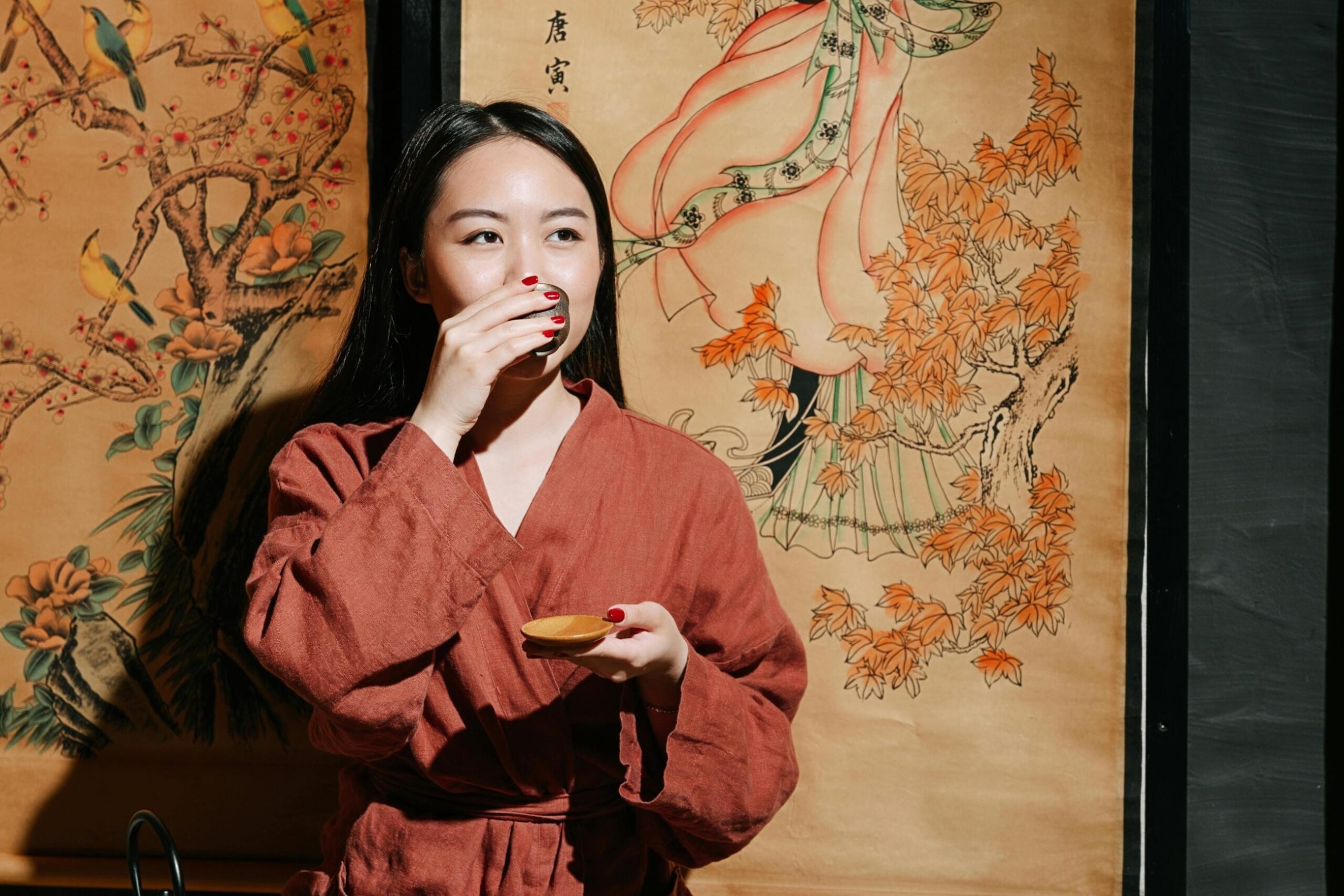 A woman wearing a textured rust-colored robe gently sips from a small cup while holding a saucer in her other hand. Behind her, traditional scroll paintings depicting birds and figures provide an artistic and culturally rich backdrop to the scene.