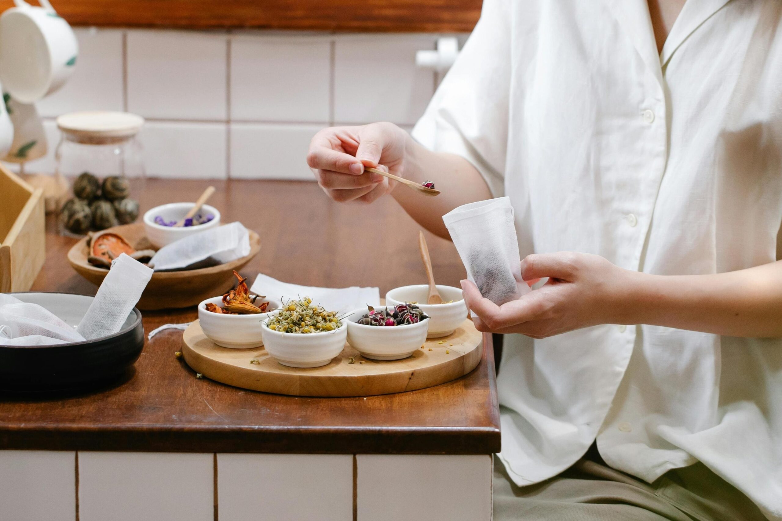 A person wearing a white shirt is shown carefully filling a tea bag with dried ingredients using a small wooden spoon. The wooden surface before them holds several small bowls containing various herbal elements, such as chamomile flowers and dried rosebuds, ready for blending.