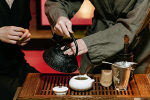A person wearing a grey robe carefully pours hot water from a black, textured cast-iron kettle into a small white teapot during a traditional tea ceremony. The ritual takes place on a slotted wooden tray adorned with various tea accessories, including a glass pitcher, strainer, and bamboo tools, while another participant sits nearby.