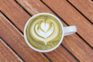 This top-down view captures a white ceramic mug filled with a matcha latte, showcasing intricate heart-shaped latte art formed in the foam. The drink rests on a warm, slatted wooden table, where the diagonal lines of the timber provide a rustic background for the vibrant green beverage.