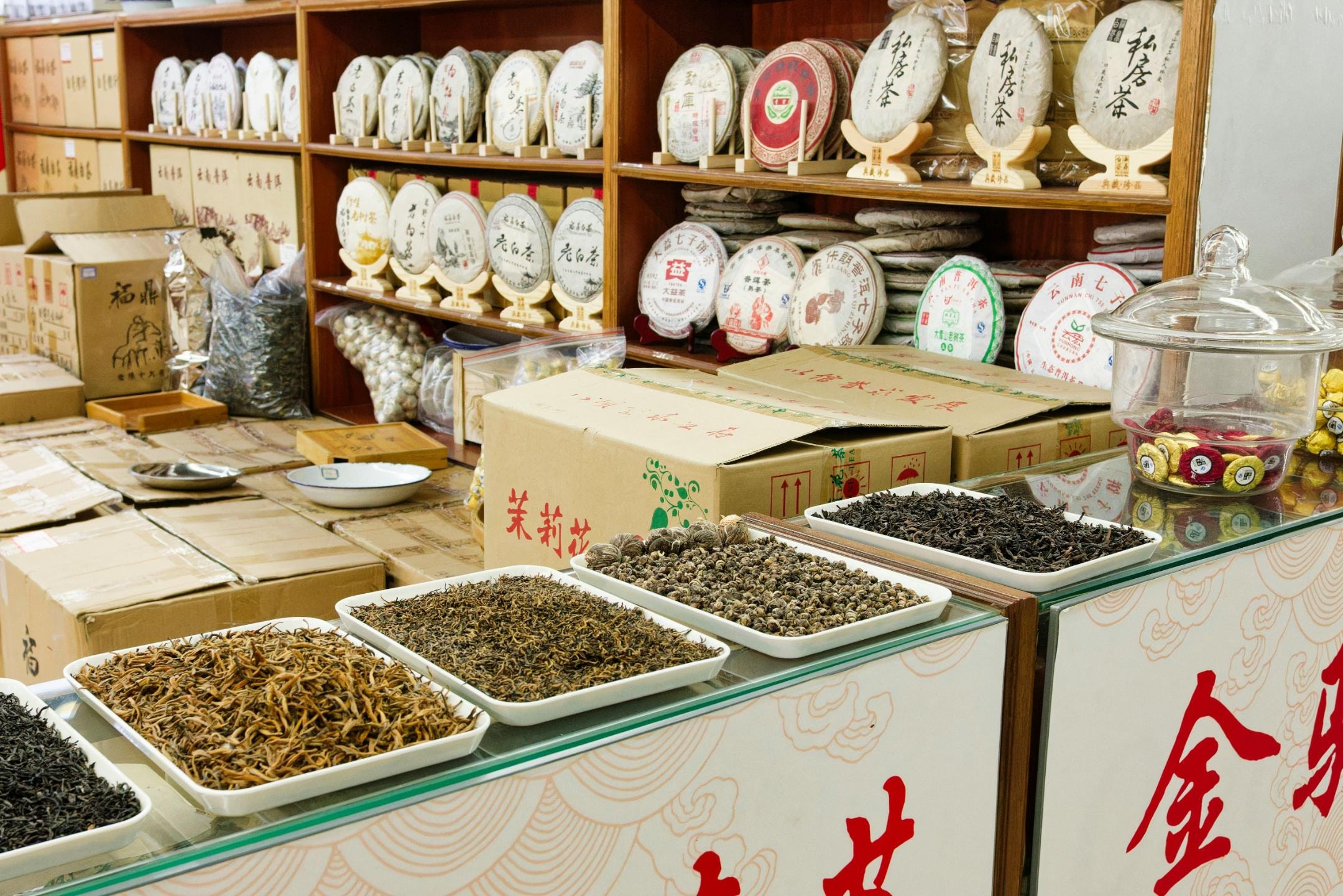 This image showcases a traditional tea shop with shelves lined with numerous wrapped tea cakes and various boxes of bulk tea. In the foreground, several trays display different varieties of loose-leaf tea, including long needle-like leaves and tightly rolled pearls, ready for selection.
