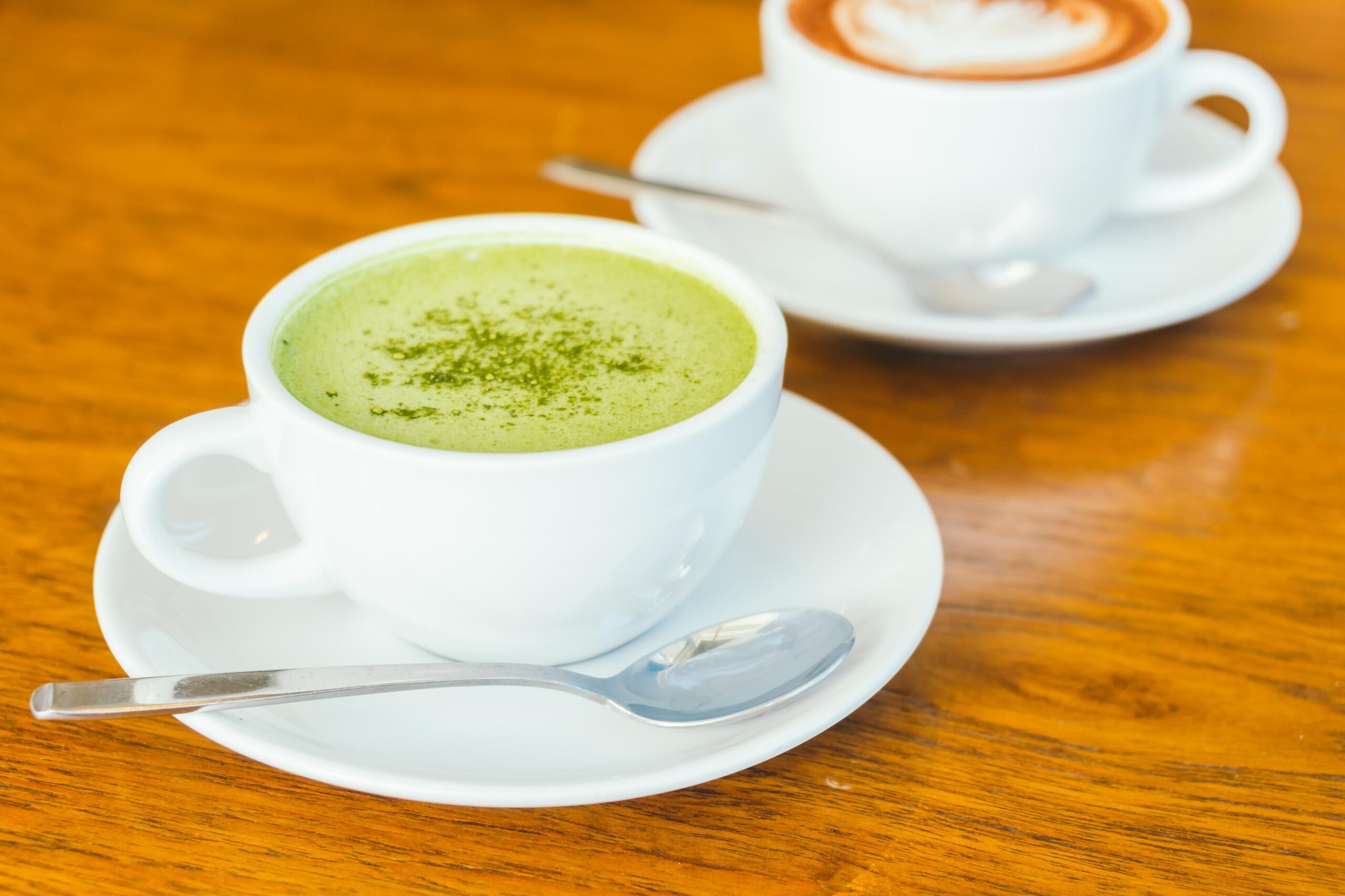 A hot matcha latte dusted with green powder sits in a white cup and saucer accompanied by a silver spoon on a wooden table. In the background, a second white cup containing coffee with latte art is visible, suggesting a shared beverage setting.
