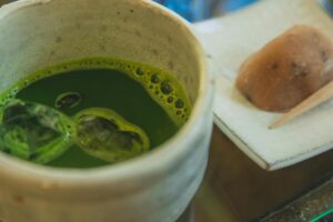 This close-up shot features a rustic ceramic cup filled with iced matcha green tea, highlighting the vibrant green color and frothy bubbles on the surface. In the softly blurred background, a traditional Japanese sweet sits on a square plate alongside a small wooden pick.