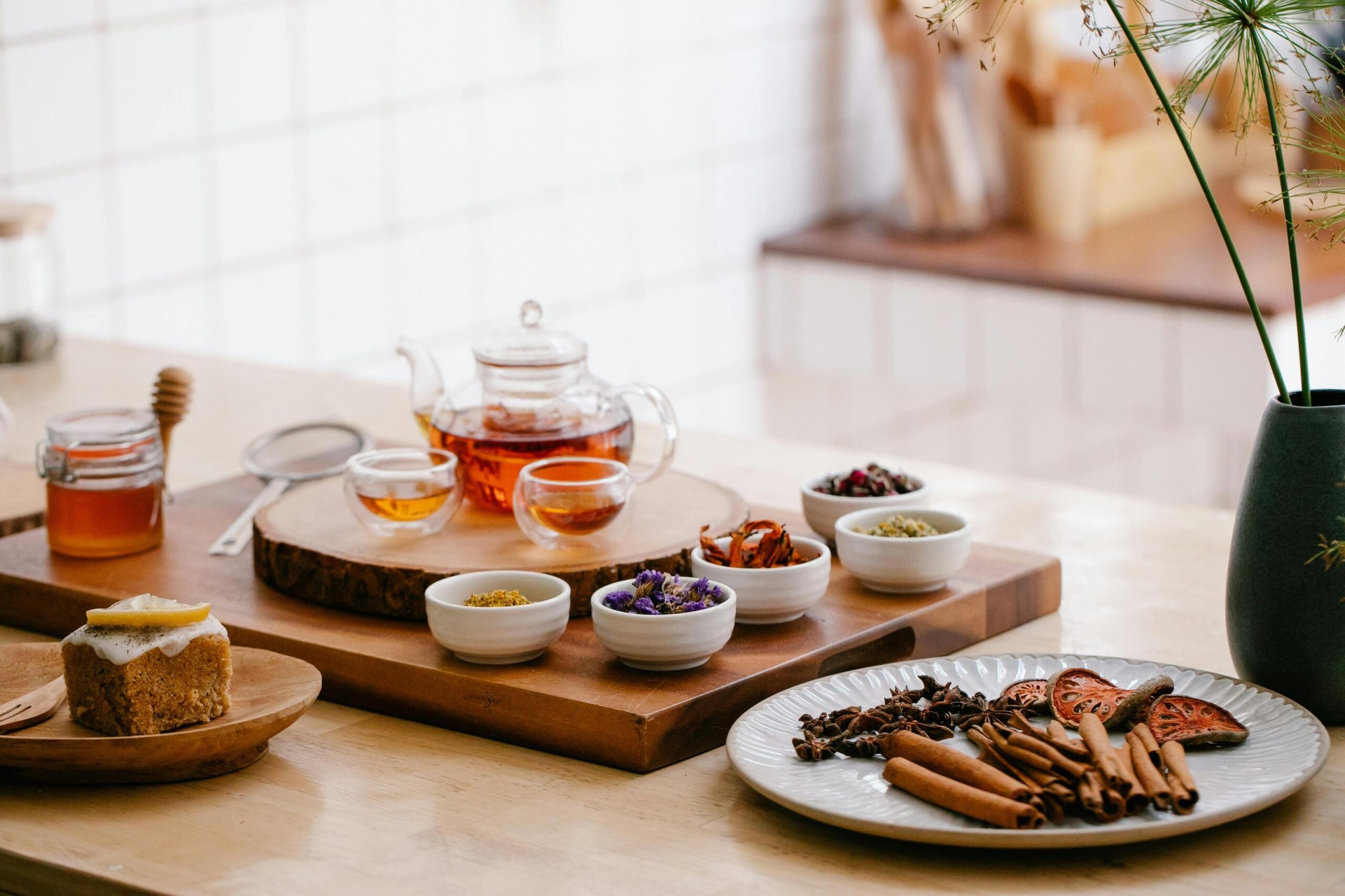 This serene kitchen scene displays a glass teapot and two small cups of amber tea set upon a rustic wooden board. Surrounding the tea service are various bowls of dried botanicals, a plate of cinnamon sticks and star anise, and a lemon-topped cake, creating a cozy atmosphere for a tea tasting.