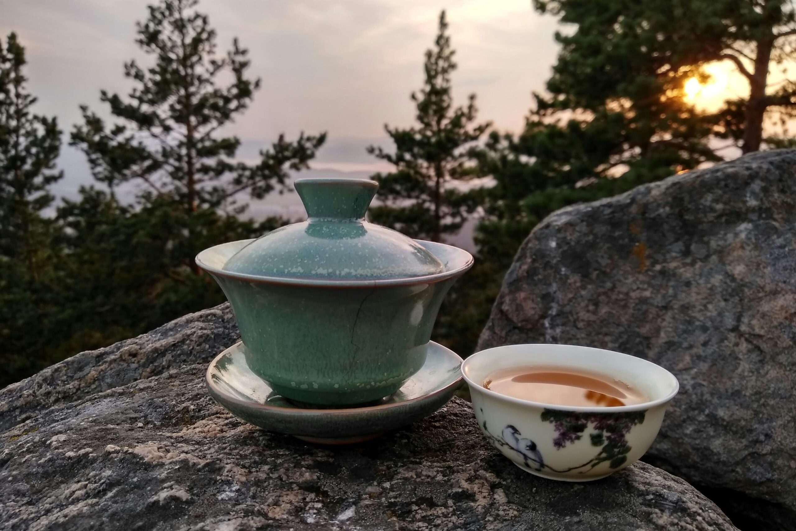 A celadon-colored gaiwan and a delicate floral tea cup filled with amber liquid rest atop a weathered grey rock. The background features a serene natural landscape with pine trees silhouetted against a soft sunset, creating a peaceful outdoor setting.