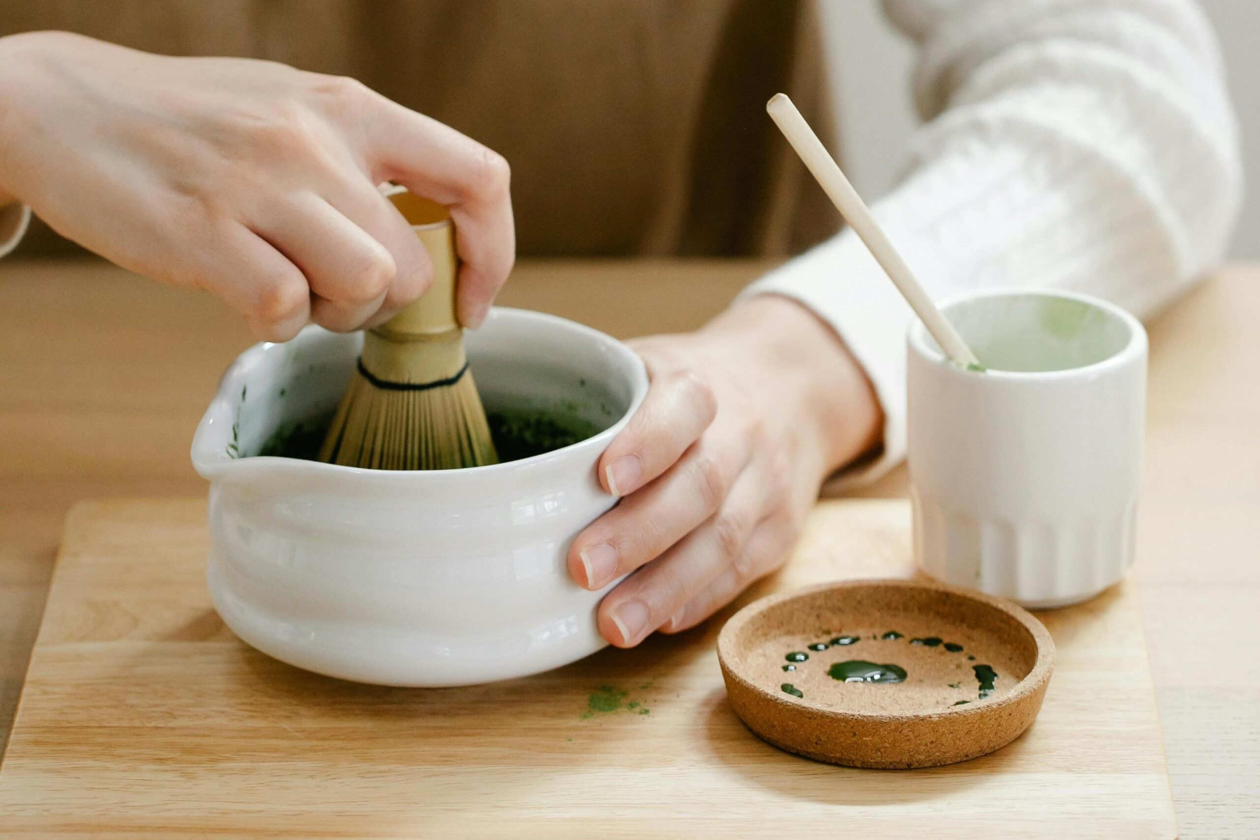This still life composition highlights a contrast of colors with a teal bowl of orange glutinous rice balls and a purple dish of green ones arranged on a wooden paddle. Beside a grey cloth napkin, a white ceramic spoon displays a single green dumpling, showcasing the glossy texture of the dessert against the dark surface.