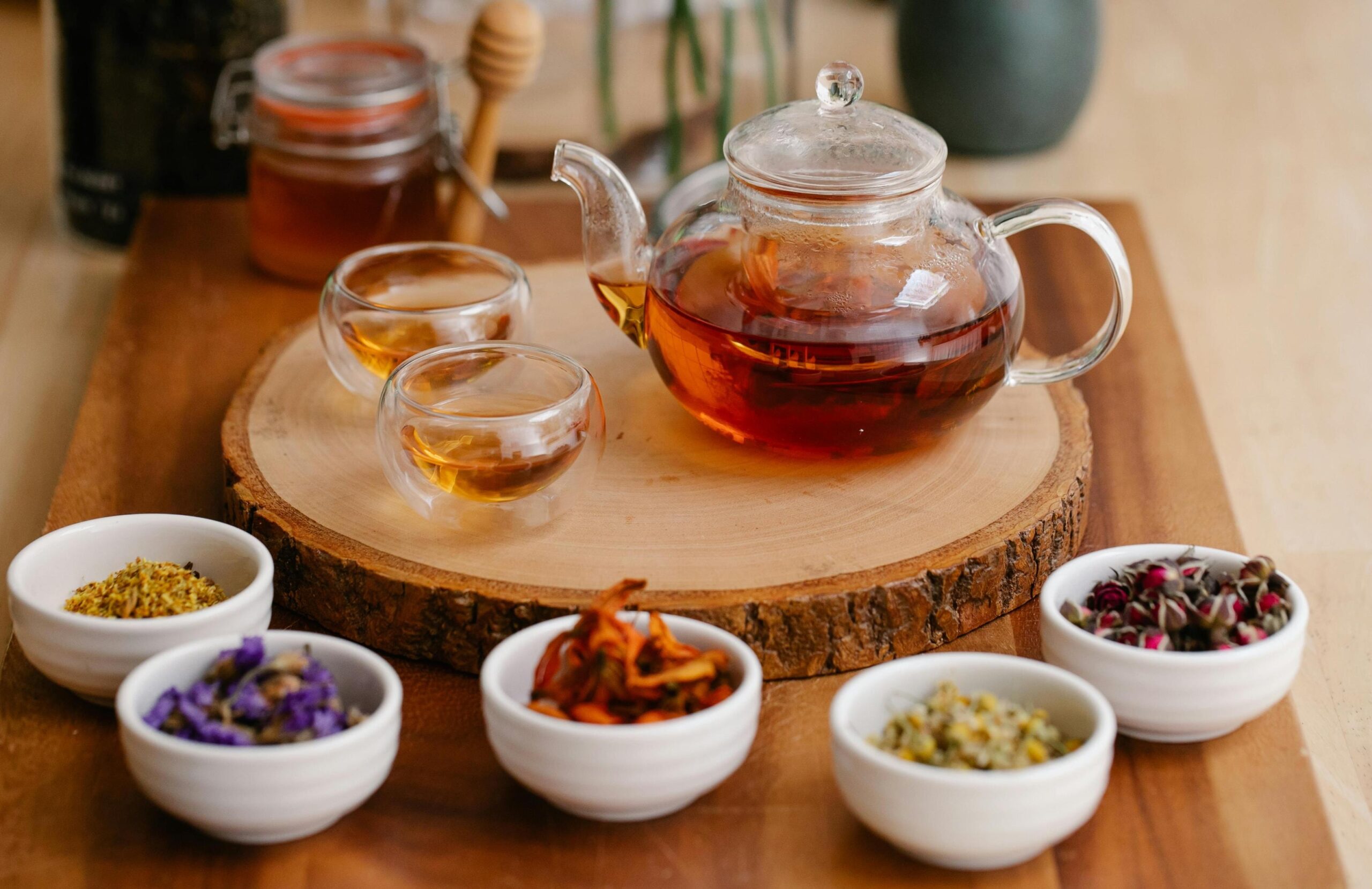 This image features a clear glass teapot and two double-walled cups filled with amber tea, all resting on a rustic, round wooden slice. Surrounding this central arrangement are five small white bowls containing various dried floral ingredients, while a jar of honey with a wooden dipper sits in the background.