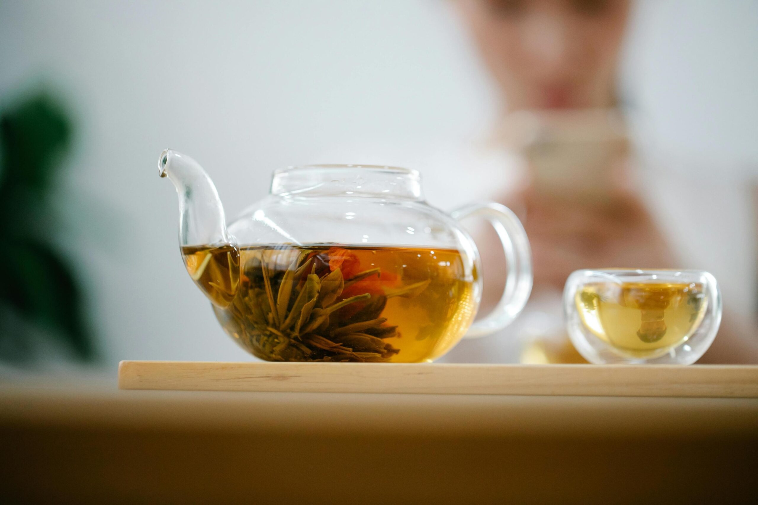 A clear glass teapot showcasing a submerged blooming flower sits beside a small glass cup of golden tea on a wooden serving board. In the blurred background, a person holds a cup to their lips, suggesting a quiet moment of enjoyment.