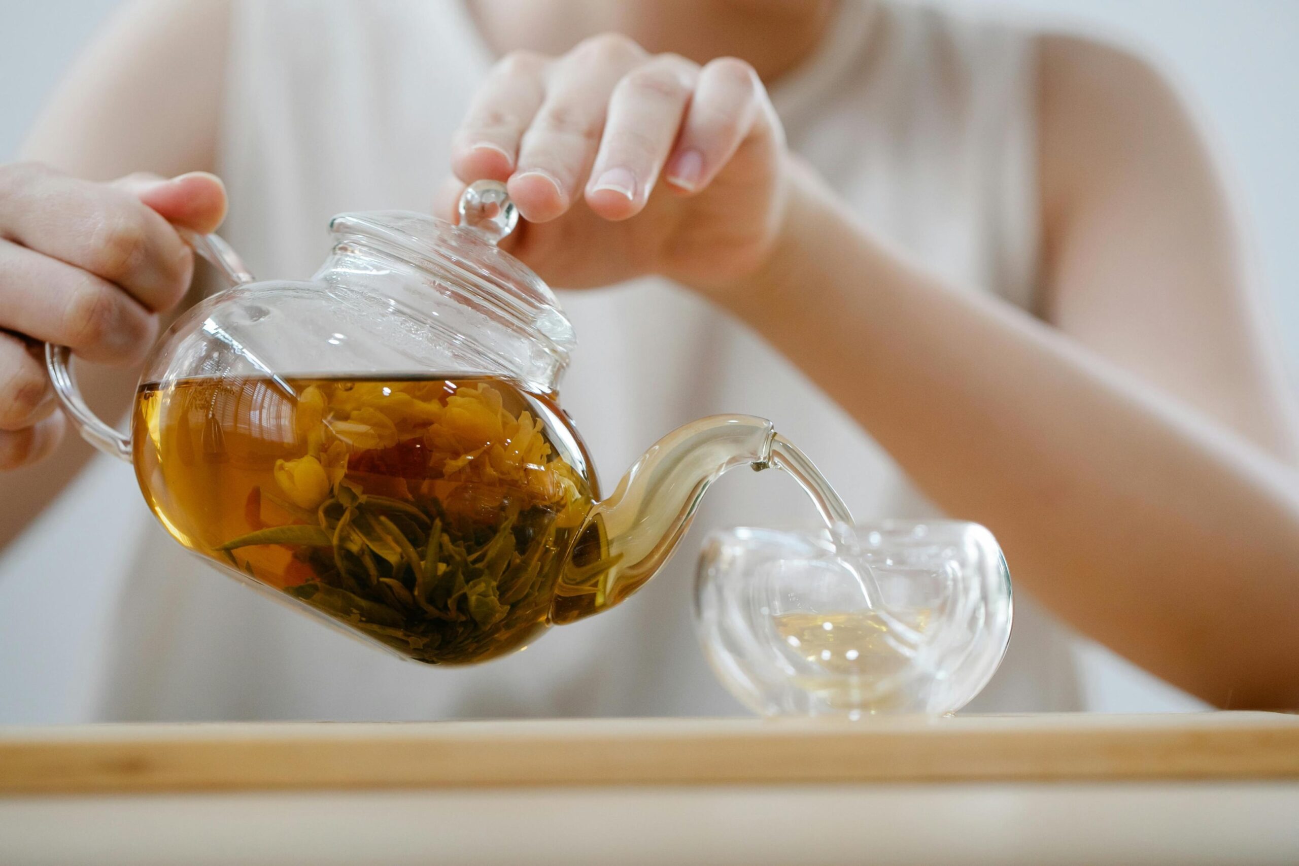 A person carefully pours amber tea from a clear glass teapot into a small, double-walled glass cup on a wooden surface. Inside the pot, a blooming tea flower is fully unfurled, adding texture and visual beauty to the golden liquid.