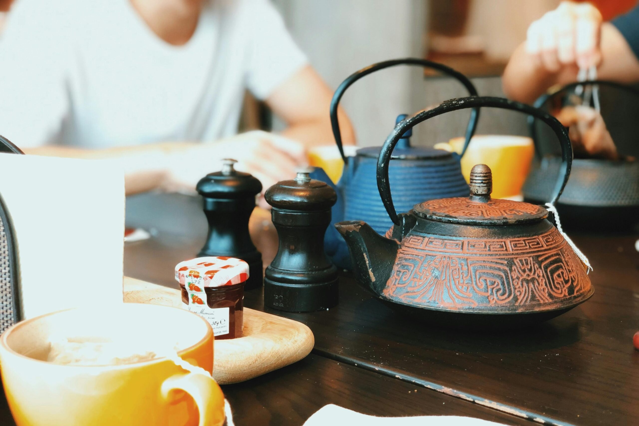 A rustic, patterned cast-iron teapot sits prominently on a dark wooden table, accompanied by black salt and pepper shakers and a small jar of jam. In the softly blurred background, diners relax in a casual cafe setting with yellow mugs and additional teapots visible.