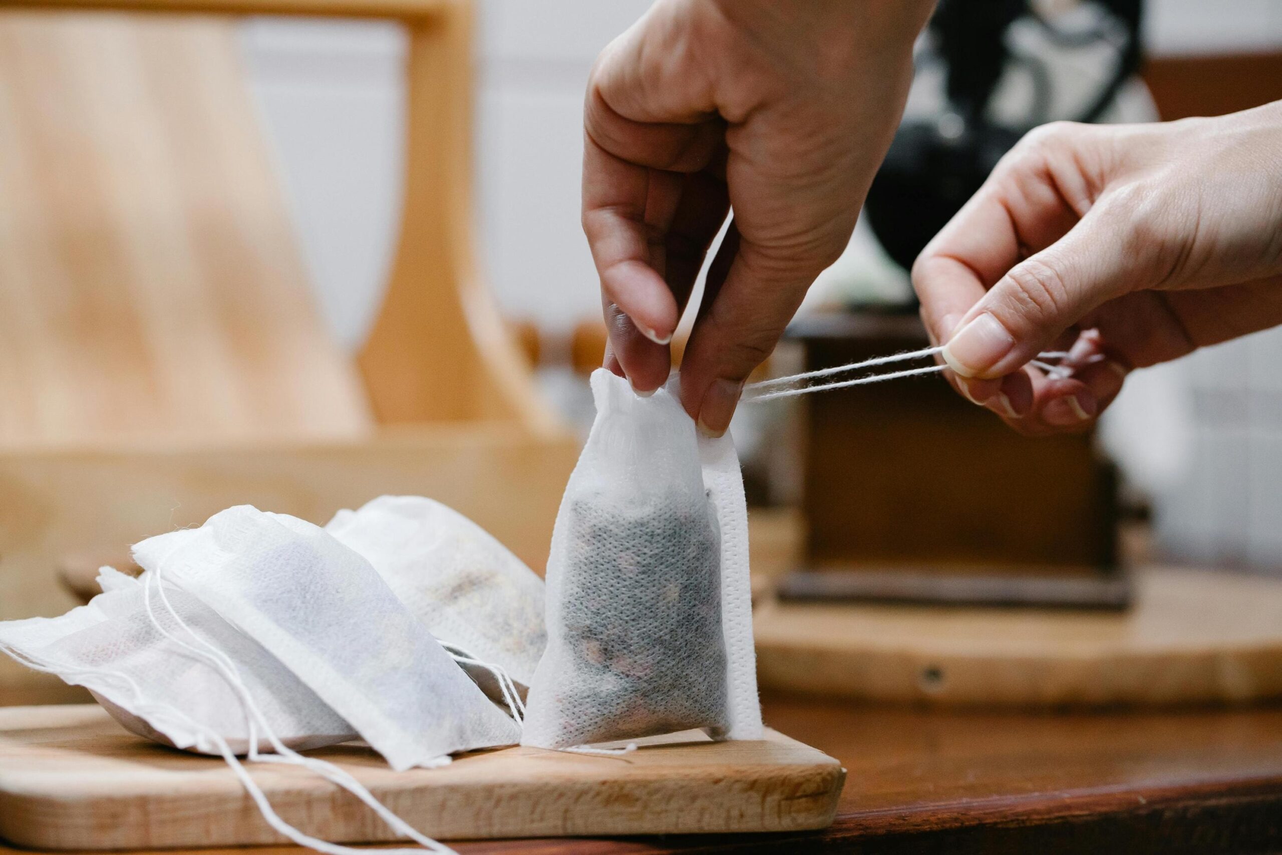 A close-up view shows a pair of hands pulling the strings to seal a white tea bag filled with dried herbs. Several other completed tea bags rest on a wooden board in the foreground, highlighting the careful preparation process.
