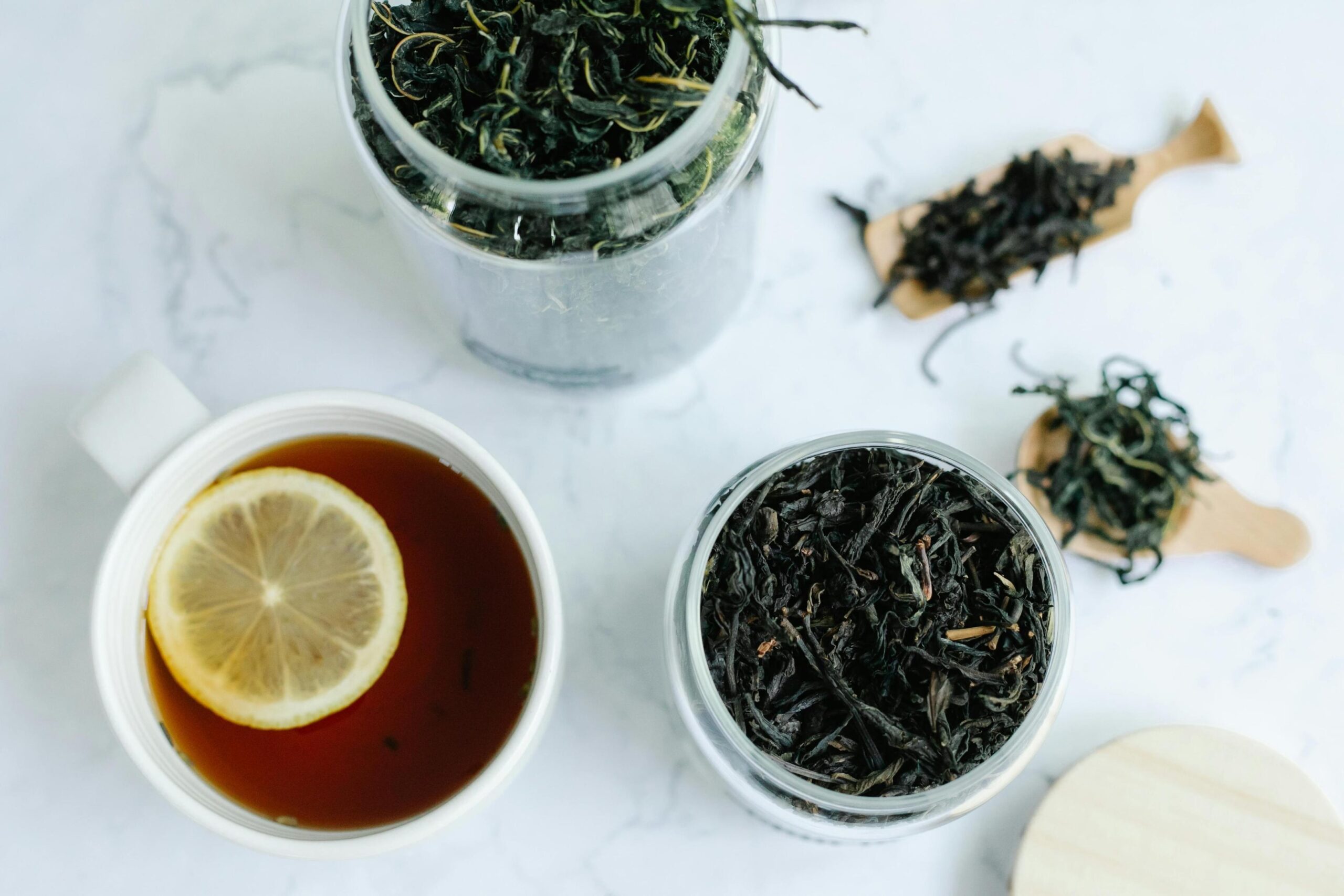 Set on a white marble-patterned surface, this overhead shot features two glass jars and small wooden scoops displaying dark, loose-leaf tea. A white ceramic mug filled with amber tea and a fresh lemon slice sits nearby, completing the refreshing arrangement.