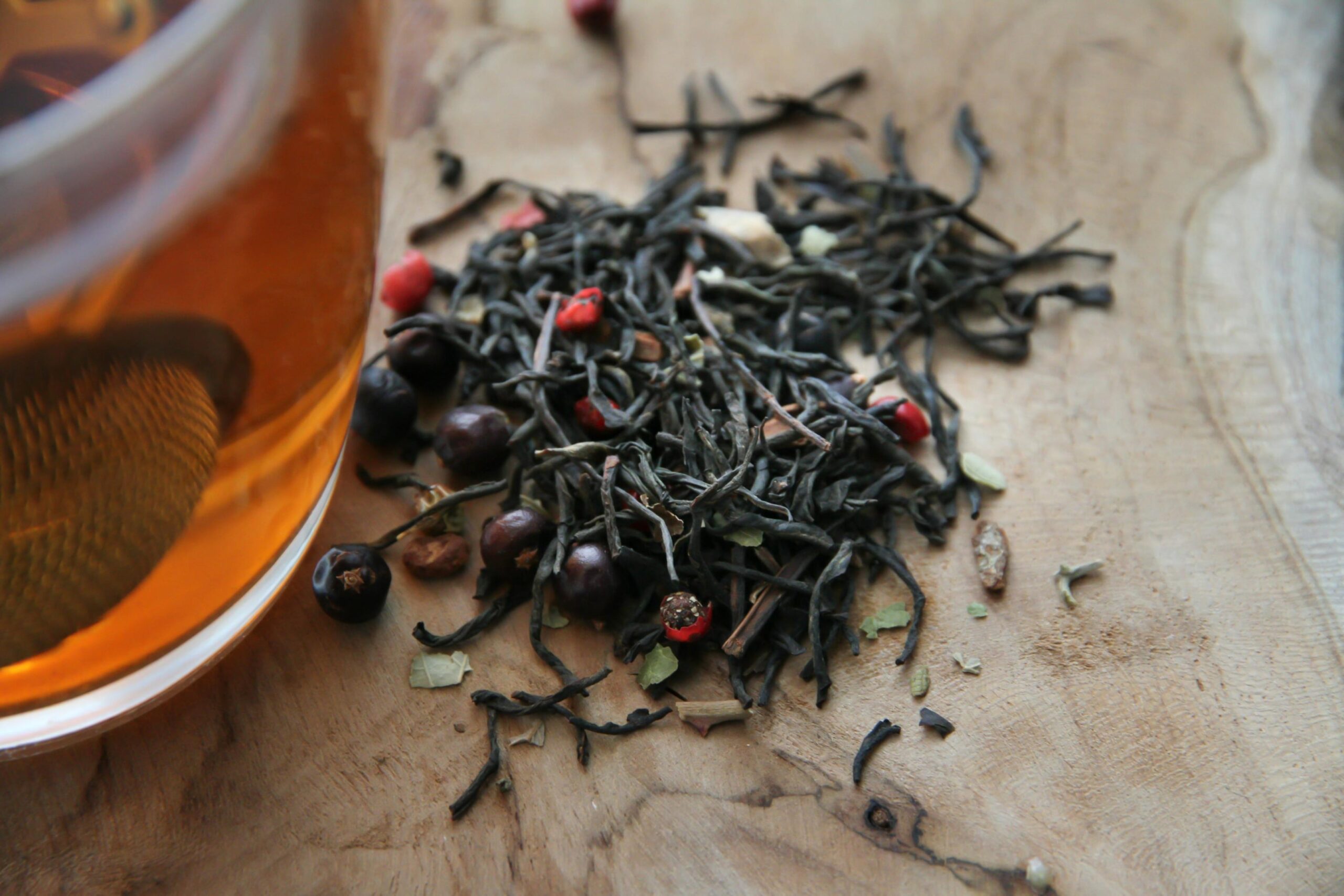 A close-up view focuses on a small pile of loose-leaf black tea mixed with dried red and dark berries resting on a light wooden surface. Partially visible on the left, a glass cup contains brewed amber liquid with a metal mesh infuser submerged, suggesting the tea is freshly prepared.