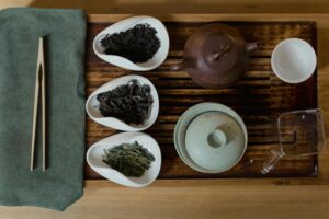 This overhead shot displays an organized traditional tea ceremony set on a wooden tray, featuring three white scoops holding different varieties of loose-leaf tea. The arrangement includes a reddish-brown clay teapot, a pale green gaiwan, a glass pitcher, and wooden tongs resting on a folded green cloth.