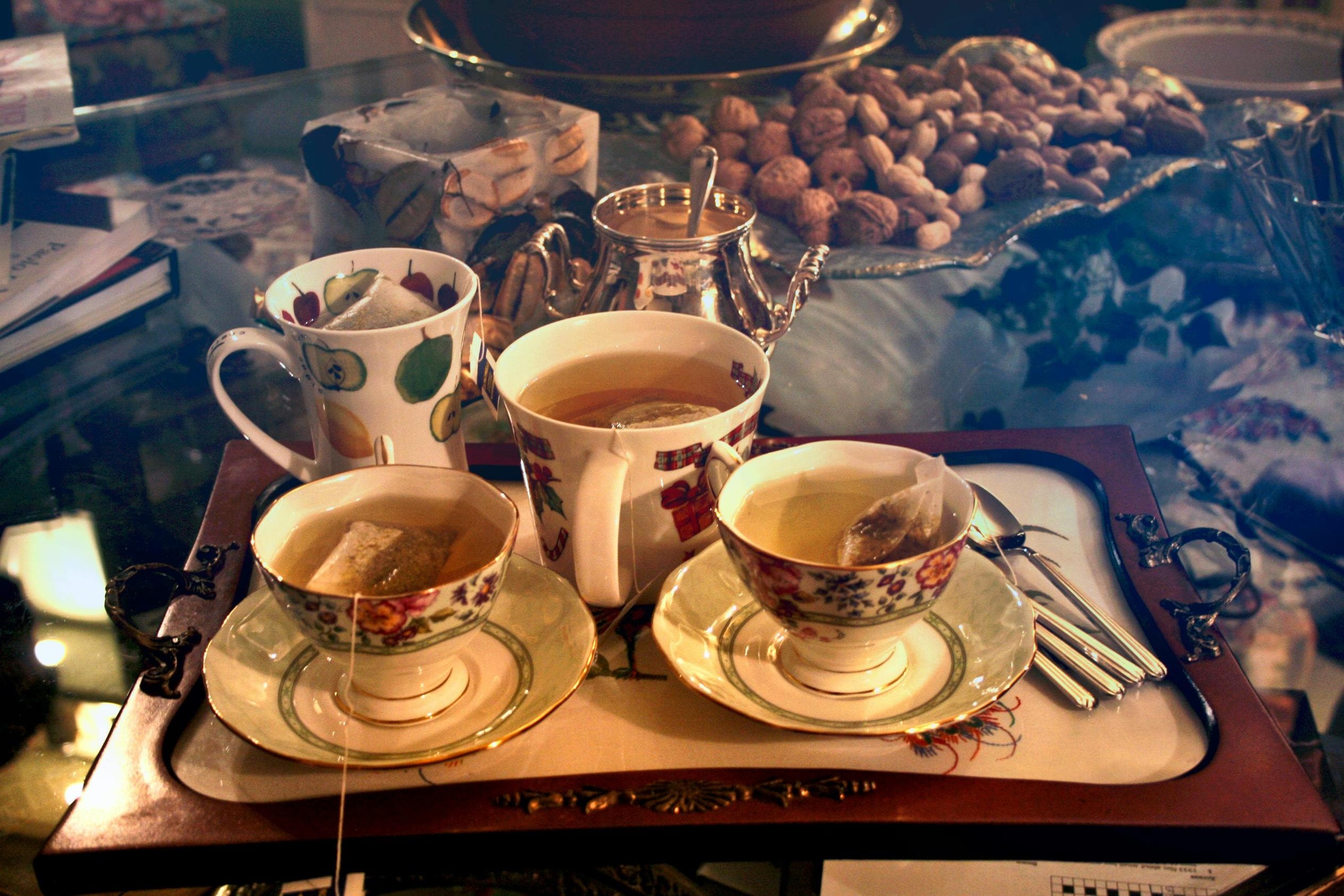 A wooden tray displays an eclectic assortment of four cups, including two delicate floral teacups and two patterned mugs, all with teabags steeping in hot water. The setting is a cluttered glass table featuring a silver bowl, a large pile of nuts in their shells, and books, evoking a cozy, domestic atmosphere.