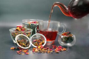 dark amber tea is poured from a glass teapot into a matching clear mug, surrounded by several glass bowls and mesh strainers filled with colorful loose-leaf tea. The vibrant blend of dried petals and herbs is scattered across the dark surface, highlighting the rich floral and botanical ingredients.