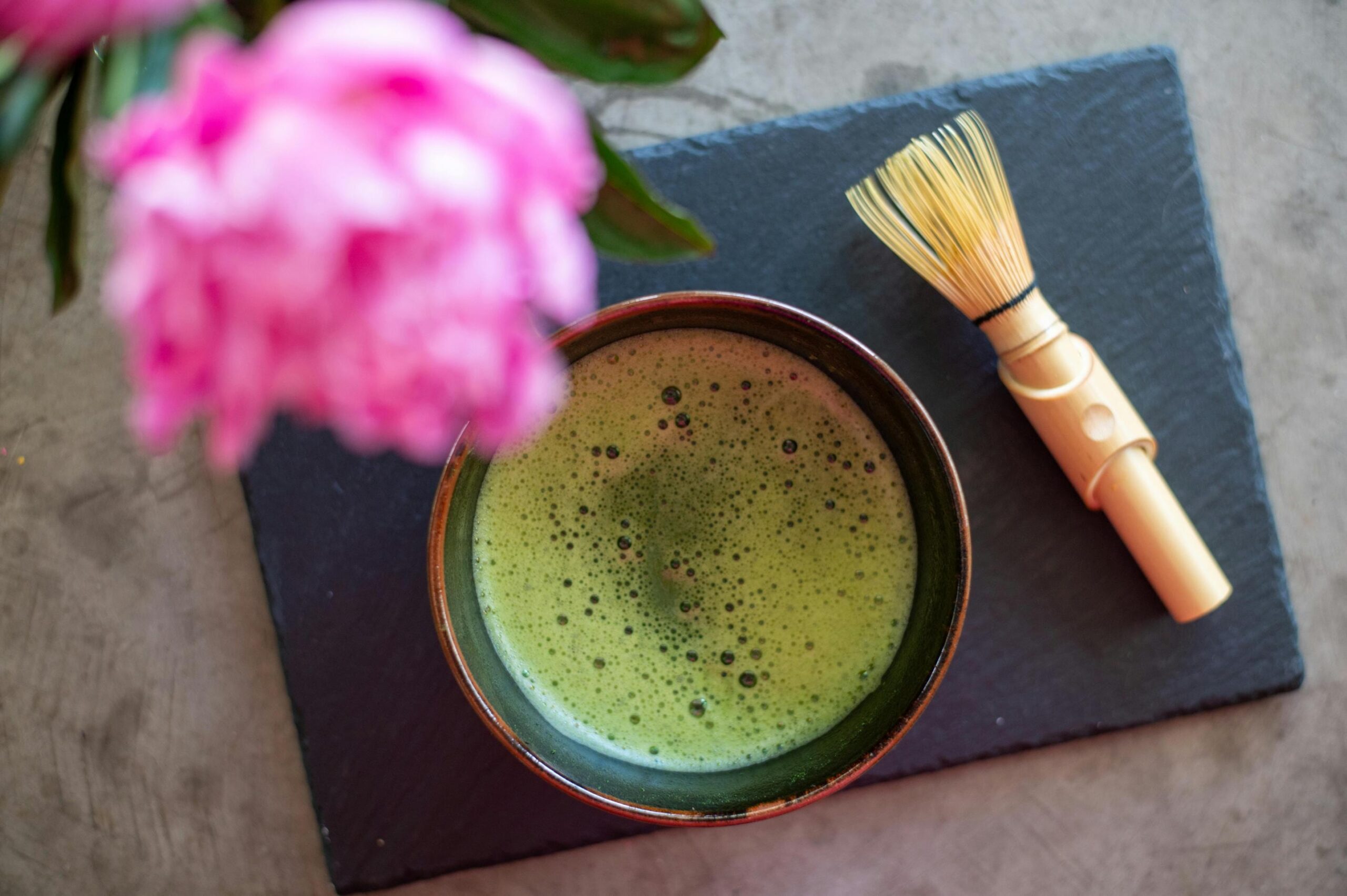 A top-down view captures a frothy bowl of green matcha tea sitting beside a bamboo whisk on a dark slate board. Vibrant pink flowers are blurred in the foreground, framing the traditional tea setup against a grey surface.