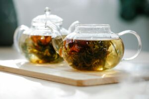 Two clear glass teapots rest on a light wooden tray, showcasing blooming tea flowers unfurling within the golden liquid. Bright natural light streams through the glass, illuminating the amber tea and creating a warm, serene atmosphere.