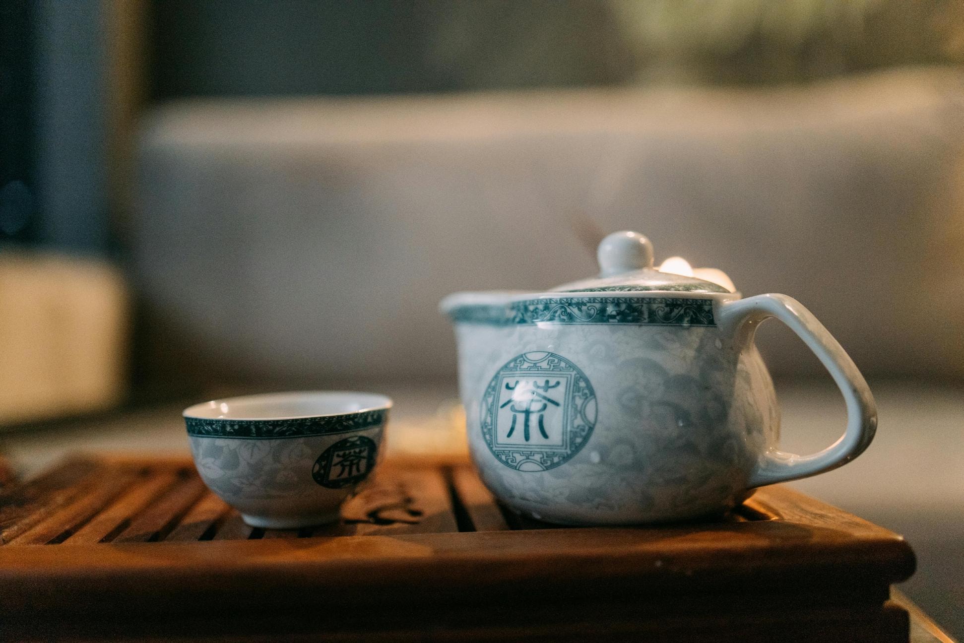 A traditional white and green porcelain tea set, featuring a teapot and matching cup adorned with Chinese characters, rests on a wooden slat tray. The image utilizes a shallow depth of field, blurring the warm, cozy background to highlight the details of the elegant ceramic ware.