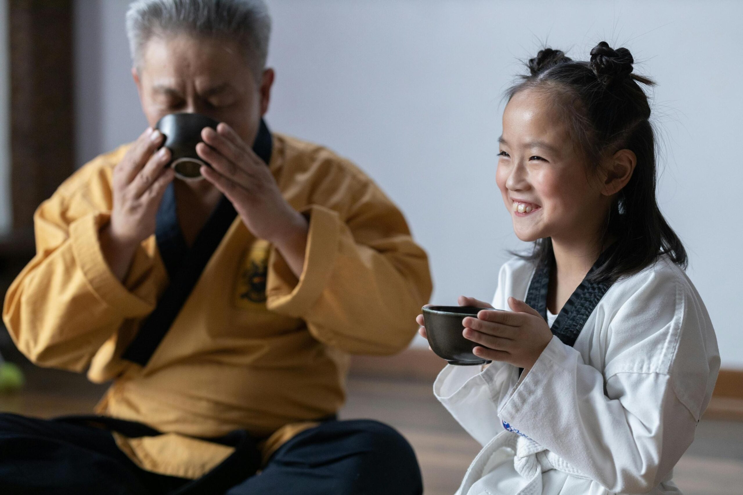 An older man and a young girl, both dressed in traditional martial arts uniforms, sit together on a wooden floor sharing a quiet moment with small cups of tea. The girl smiles broadly while holding her cup with both hands, contrasting with the man who is focused on sipping his drink in the background.