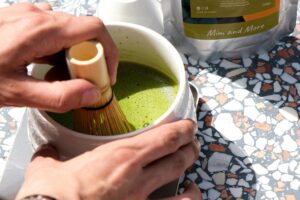 A person uses a traditional bamboo whisk to froth bright green matcha tea inside a white ceramic bowl. The preparation takes place on a sunlit terrazzo table, with a pouch of the tea powder visible in the background.