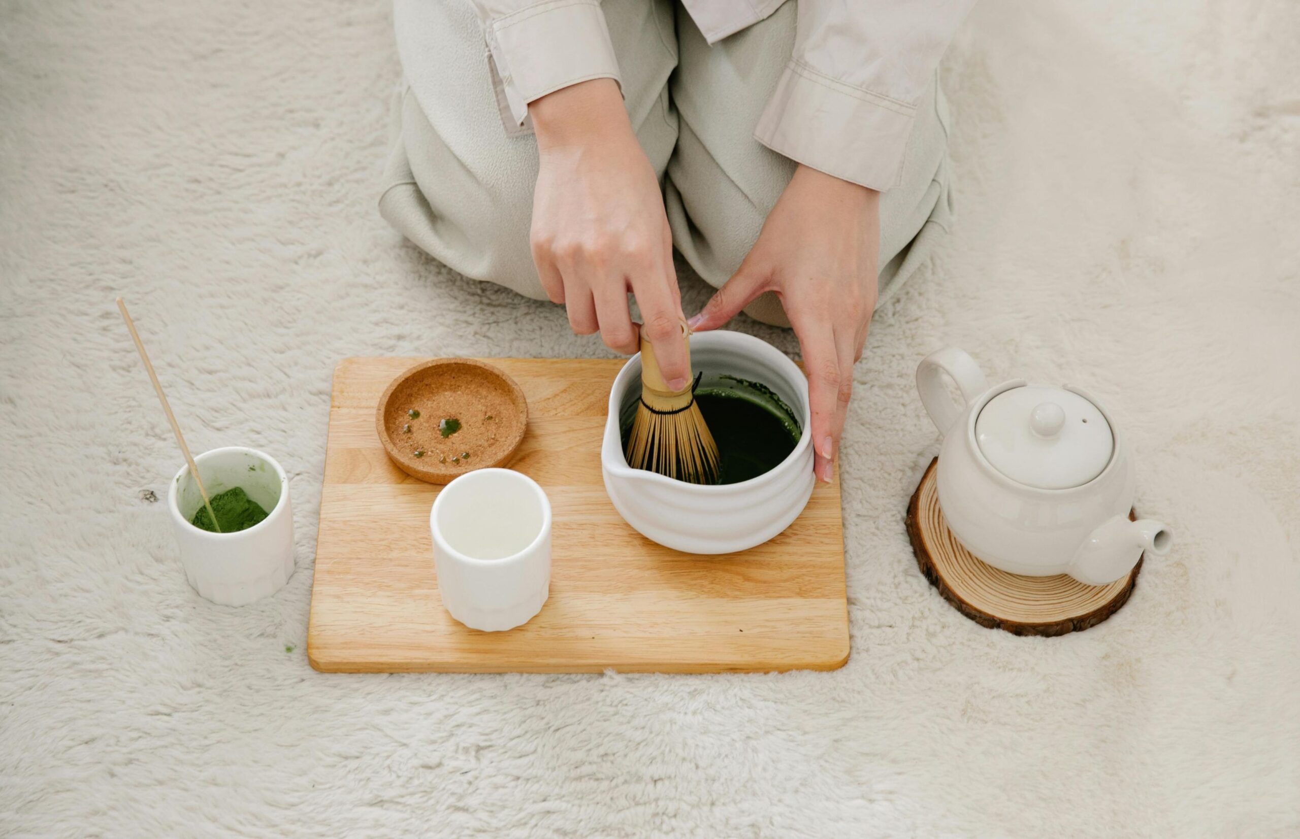 Captured from above, a person kneels on a plush white rug while whisking green matcha in a white bowl using a traditional bamboo whisk. The serene scene includes a light wooden board holding tea essentials, flanked by a matching white teapot resting on a rustic wood slice.