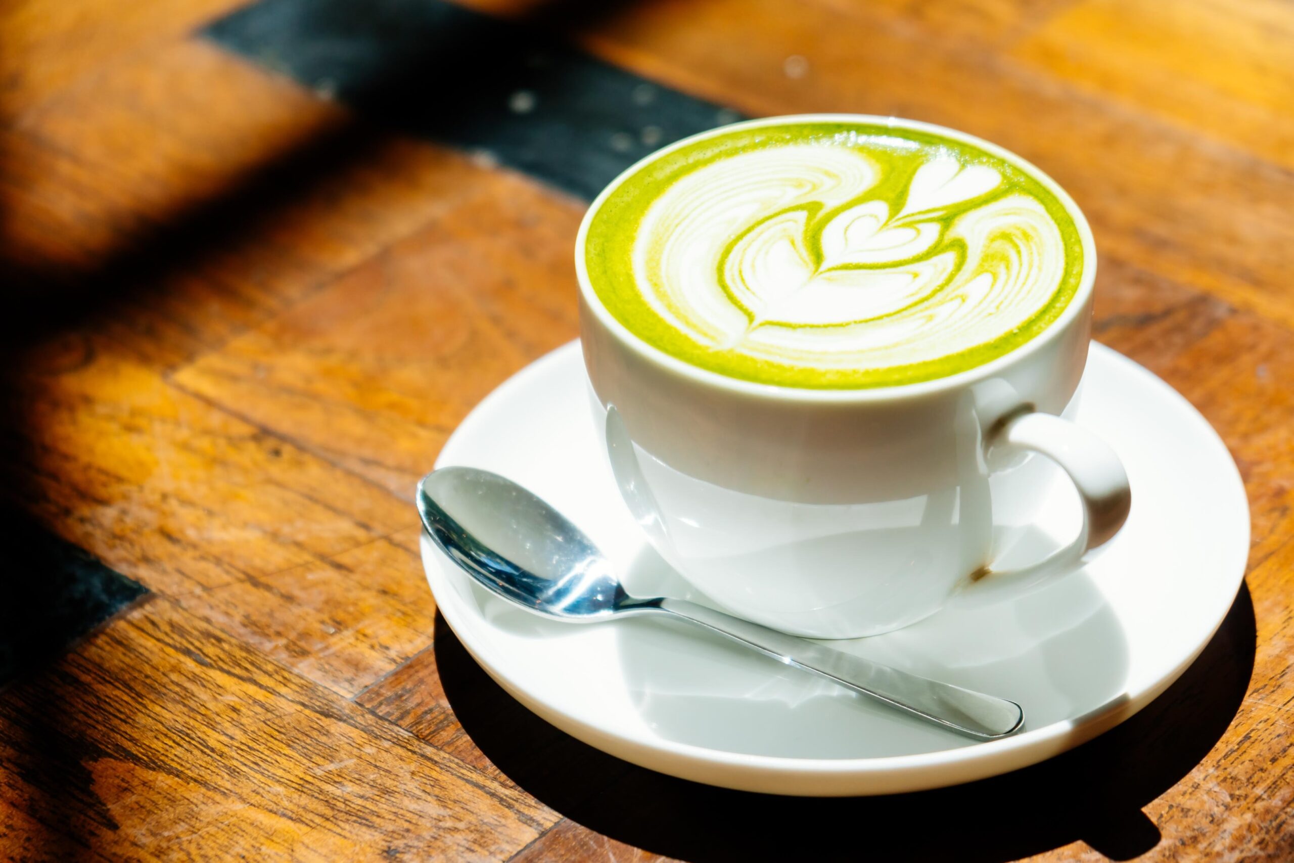 A white cup filled with a hot matcha latte features intricate heart-shaped foam art on its vibrant green surface. It rests on a matching saucer alongside a silver spoon, sitting on a sunlit wooden table that casts sharp shadows.