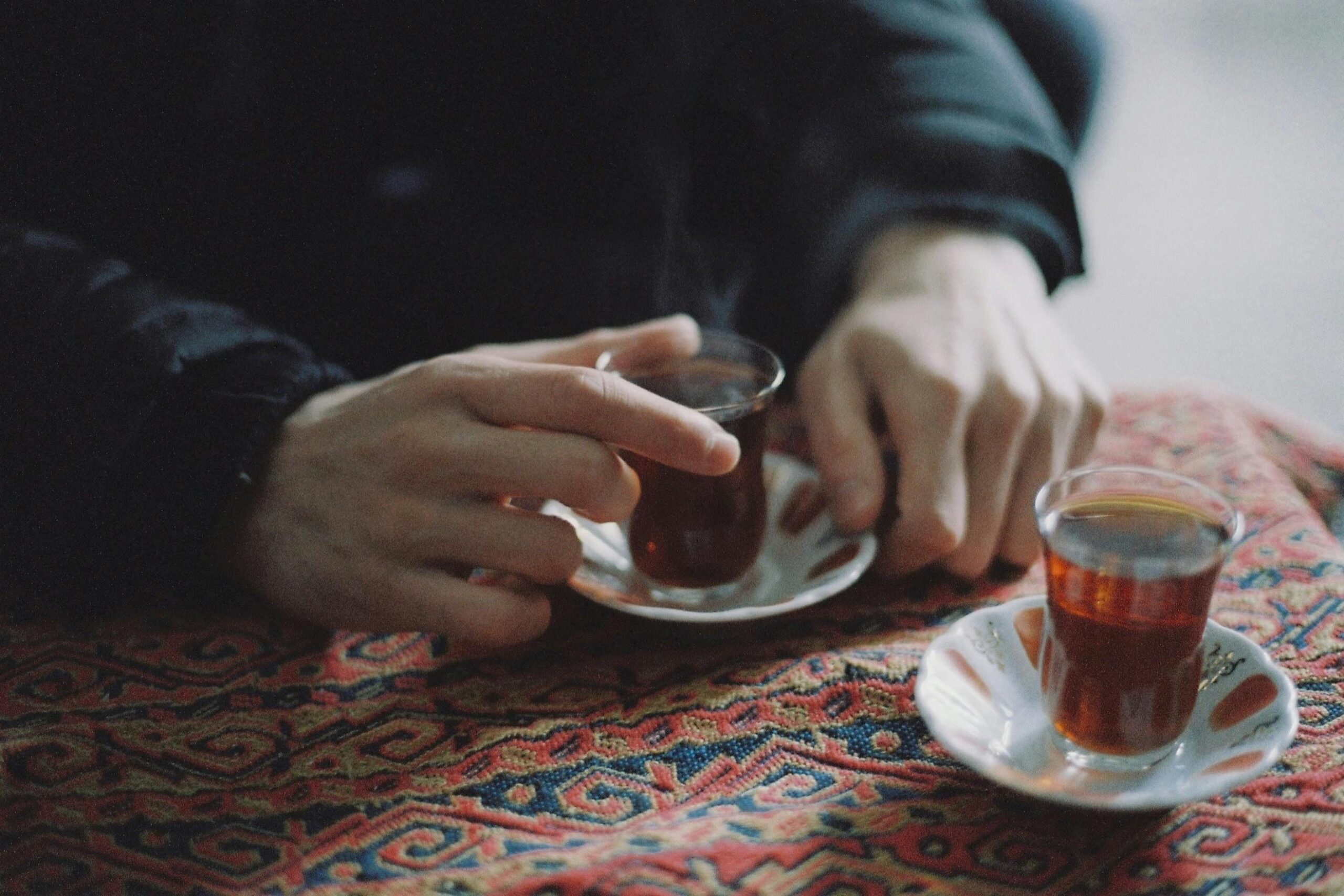 A person's hands gently grasp a small, hourglass-shaped glass of dark tea resting on a vibrant, intricately patterned textile. A second glass sits nearby on a matching saucer, suggesting a shared moment over hot beverages in a culturally rich setting.