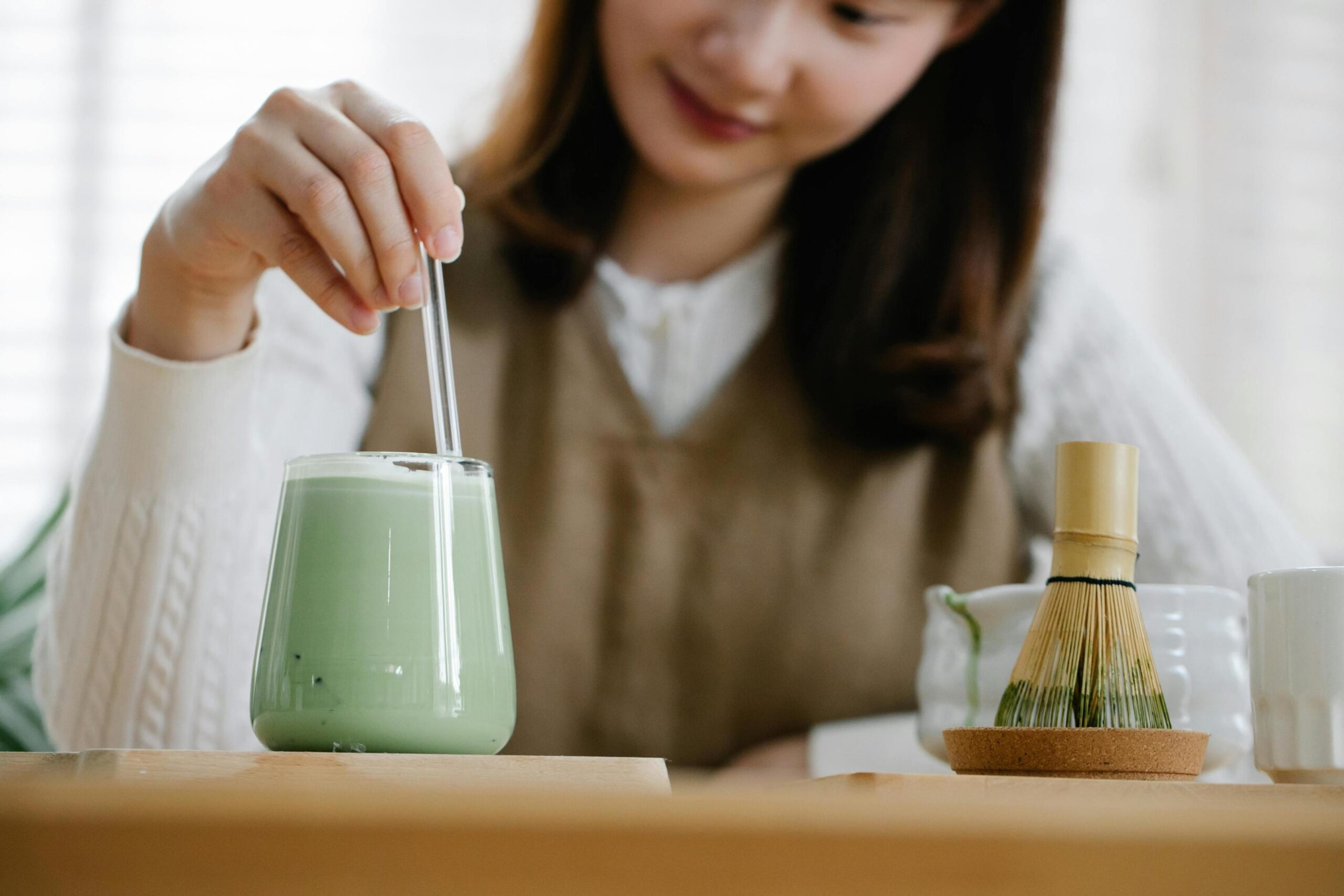 A woman gently stirs a glass of iced matcha latte with a clear straw, her focus directed at the pale green drink. To the right of the glass, a traditional bamboo whisk sits on the wooden table, highlighting the preparation of the tea.
