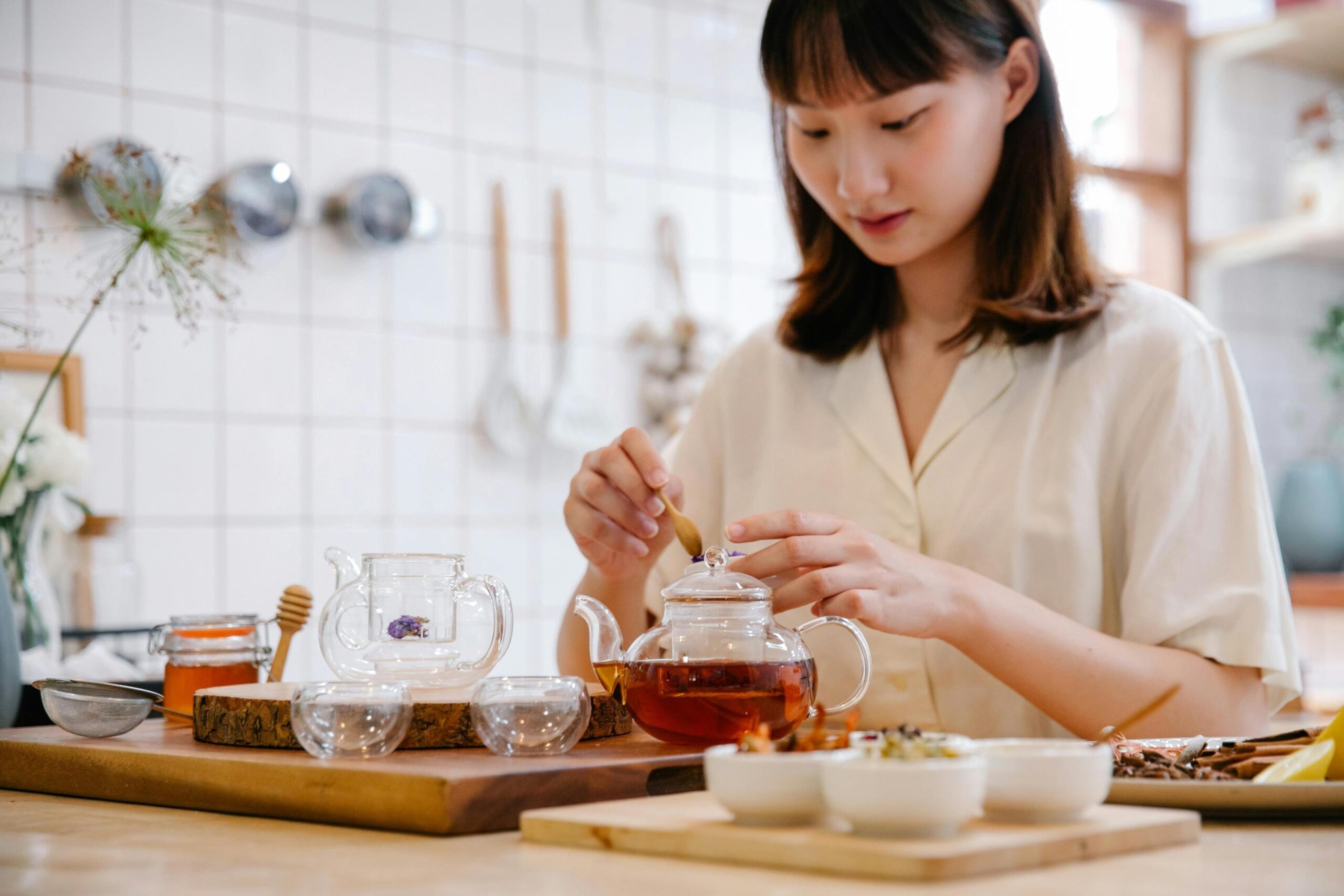 A young woman in a light-colored shirt sits at a wooden table, carefully adding ingredients to a clear glass teapot using a small wooden spoon. The bright, tiled kitchen setting features a spread of tea-making essentials, including small glass cups, a jar of honey, and bowls of dried herbs and spices.
