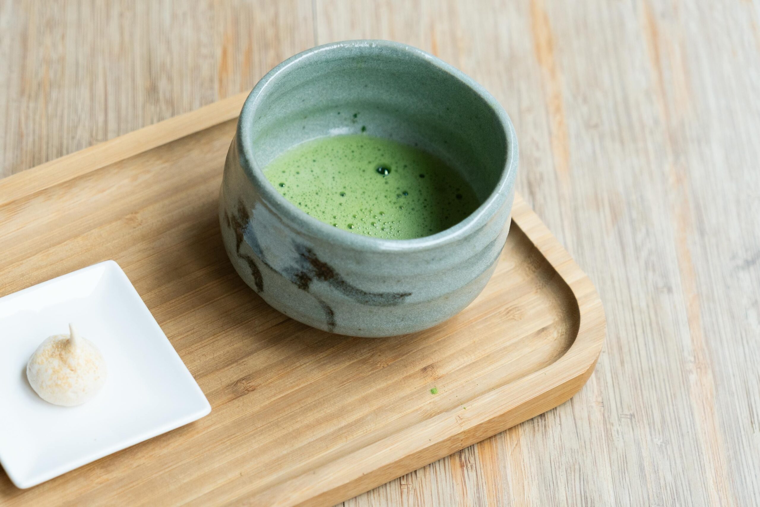 A rustic, light-blue ceramic bowl filled with frothy green matcha tea sits on a simple wooden tray. Beside the tea, a small white square plate holds a single, delicate white confection dusted with powder.