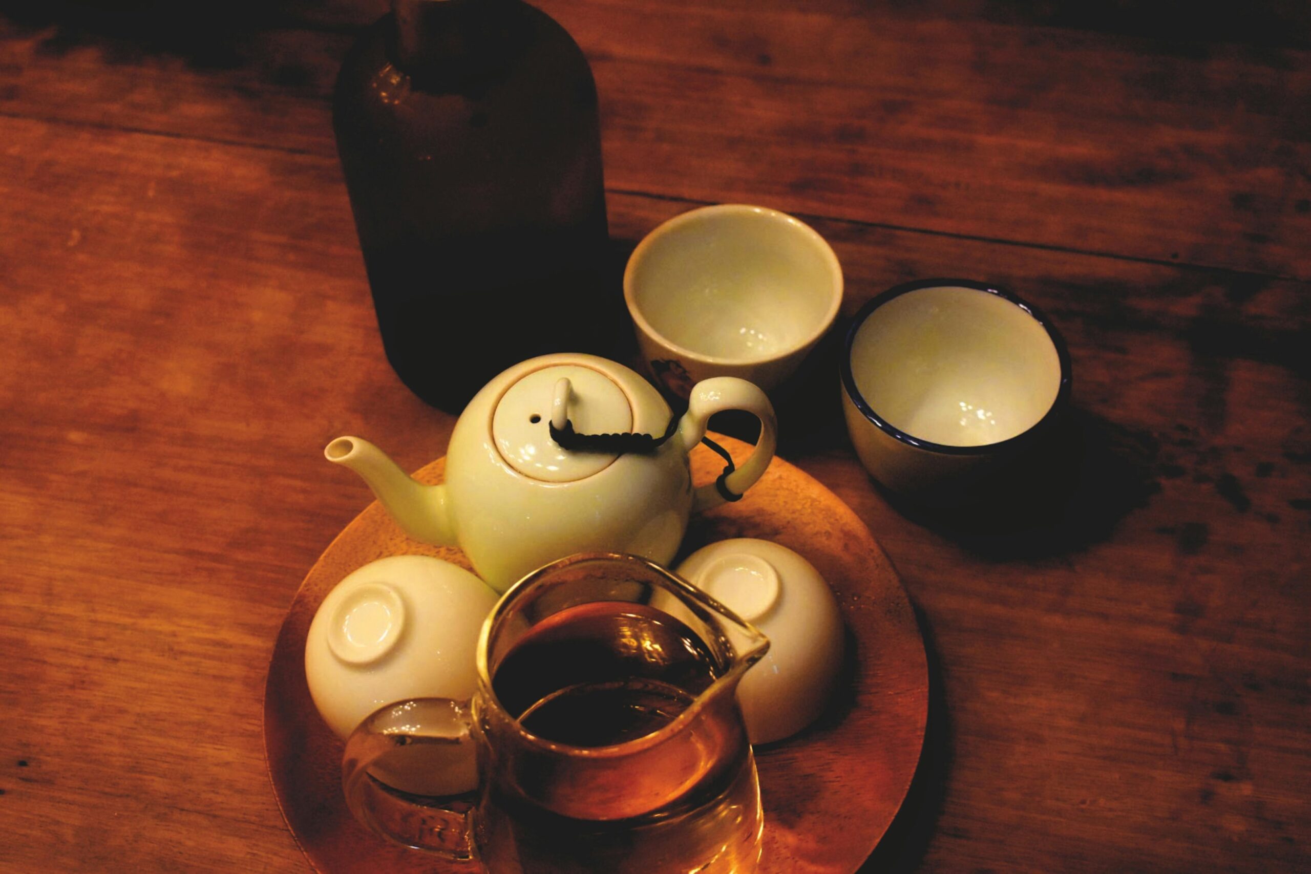 Under warm, dim lighting, a small cream-colored teapot and a glass pitcher filled with amber tea rest on a round wooden tray. Surrounding the central set on the rustic table are small ceramic cups—some upright and others overturned—along with a tall dark bottle standing in the shadows.