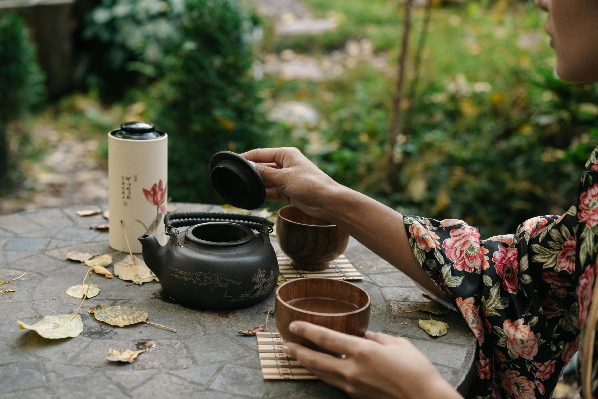  A person wearing a floral kimono sits at a stone table in an outdoor garden, holding a wooden cup while removing the lid from a dark, etched teapot. The serene setting is cluttered with fallen autumn leaves and includes a tall decorative tea canister and wooden coasters.