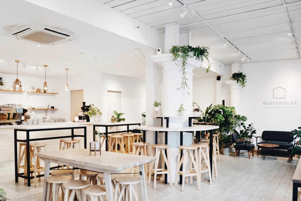  This image displays the bright and airy interior of a modern cafe called "Glasshouse," characterized by its minimalist white walls, light wood furniture, and abundant green plants. The space features various seating options including high-top tables with wooden stools and a cozy lounge area with dark sofas, all set under a clean white ceiling with industrial-style track lighting.