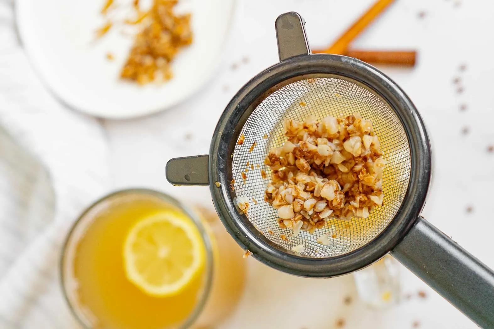 A close-up, top-down view shows a metal mesh strainer filled with soft, expanded buckwheat groats held over a glass of amber-colored tea. In the blurred background, a slice of lemon floats in another glass of tea alongside a cinnamon stick on a white surface.