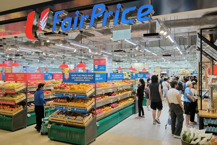 A vibrant, well-lit FairPrice supermarket in Singapore's City Square Mall features shoppers browsing through organized displays of fresh produce under modern industrial-style ceilings.