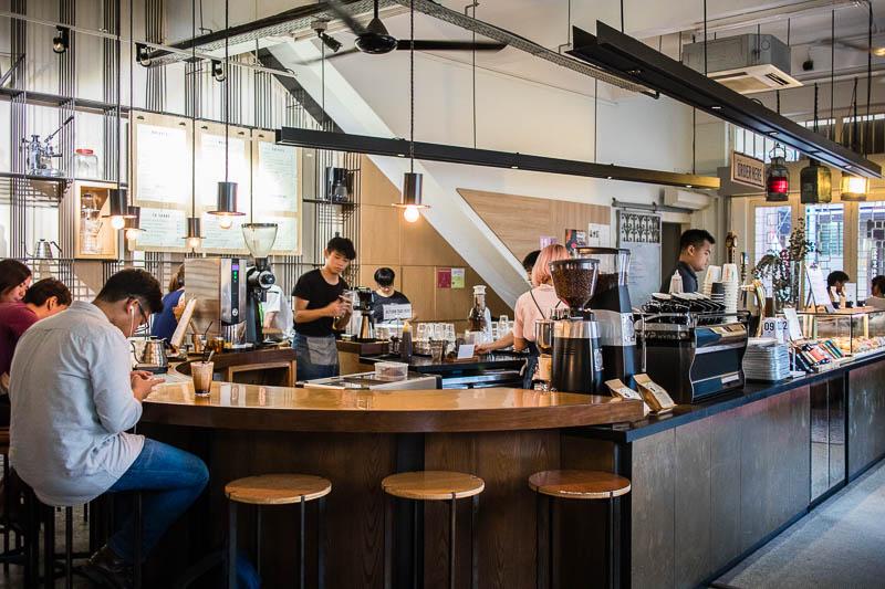 A busy, modern coffee shop features a curved wooden bar where customers sit and baristas prepare drinks behind professional espresso equipment. The interior is brightly lit with industrial-style pendant lamps and features a clean aesthetic with light wood paneling and white walls.