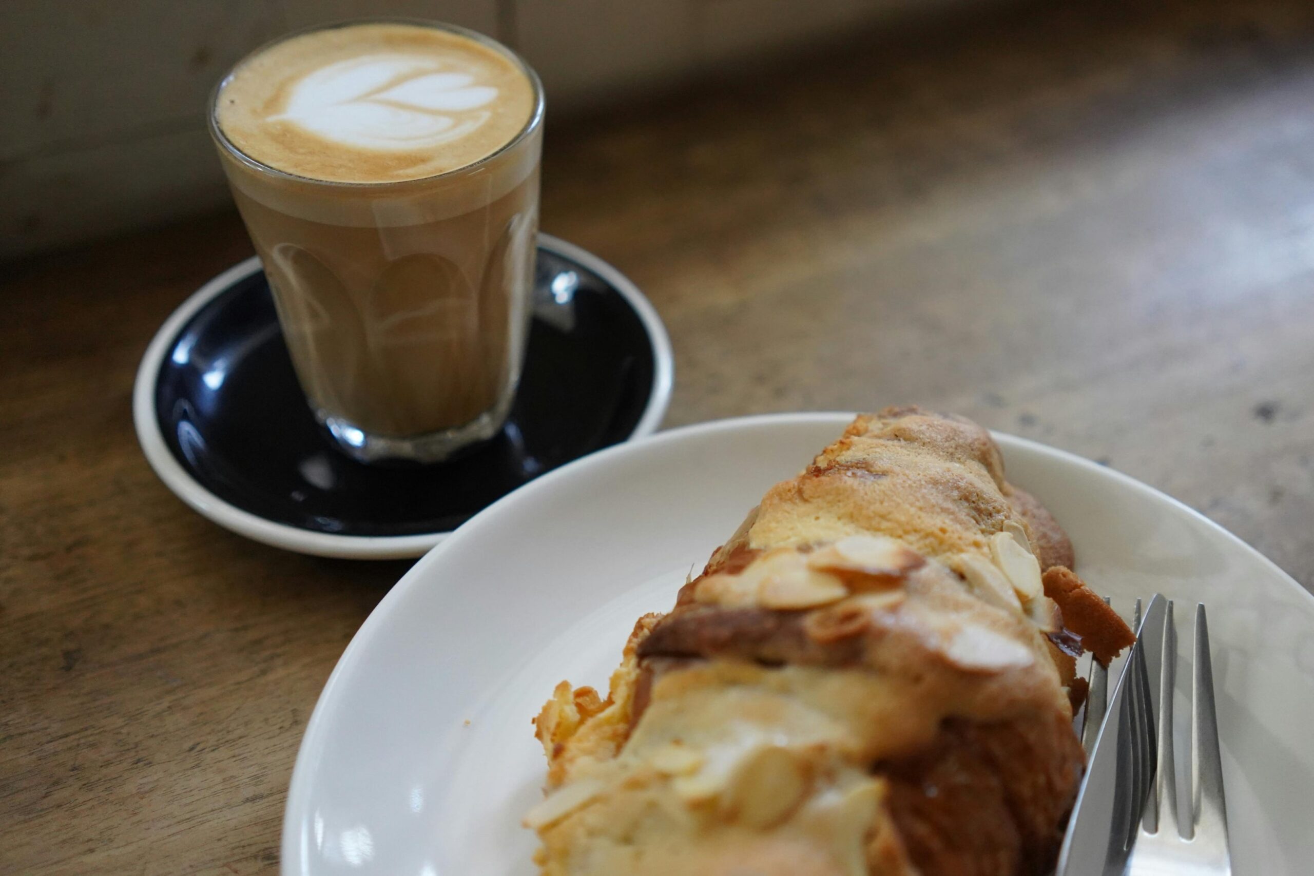  A latte with leaf-shaped latte art is served in a small glass on a dark saucer alongside a golden-brown almond croissant. Both items are presented on a rustic wooden table, with a metal fork visible on the edge of the white plate holding the pastry.