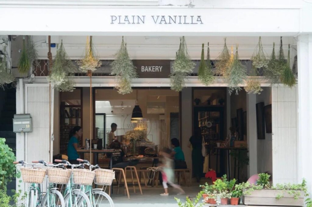 The exterior of this "Plain Vanilla" bakery features a white storefront adorned with rows of hanging dried flowers and greenery. Several mint-green bicycles with wicker baskets are parked out front, creating a charming and rustic atmosphere for customers inside.