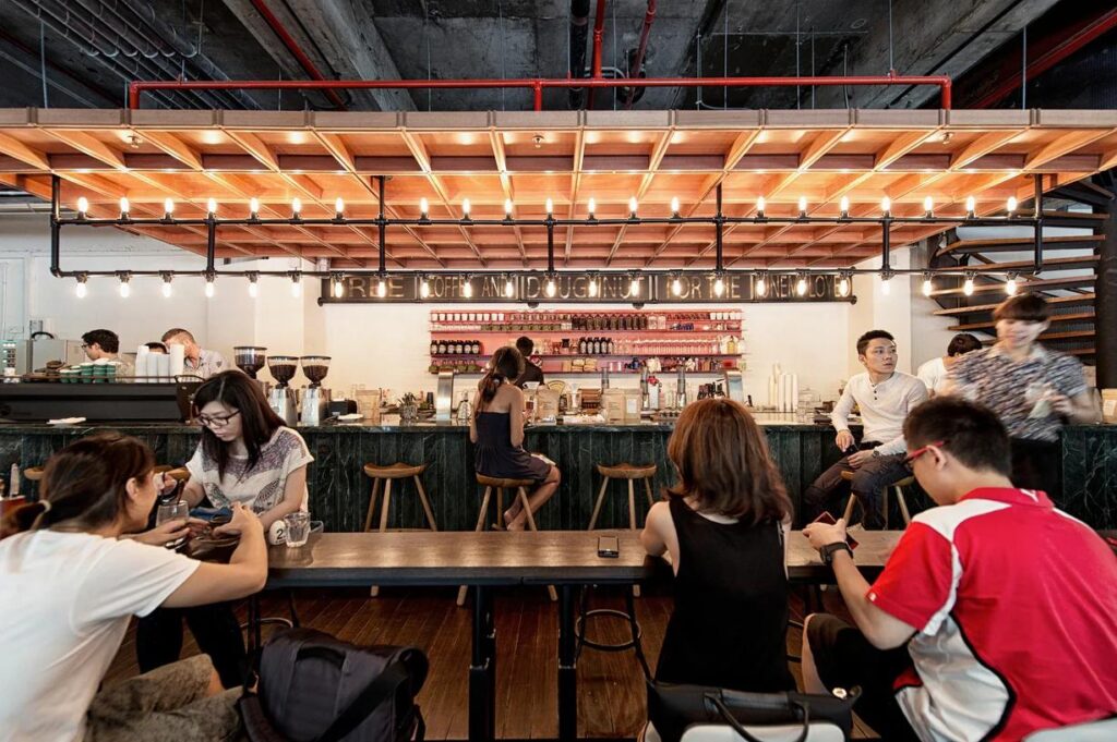 This industrial-chic cafe features an impressive overhead light installation made of black piping and warm Edison bulbs. Patrons are seated at dark communal tables and a marble-topped bar, all set against a backdrop of exposed concrete ceilings and red accent pipes.