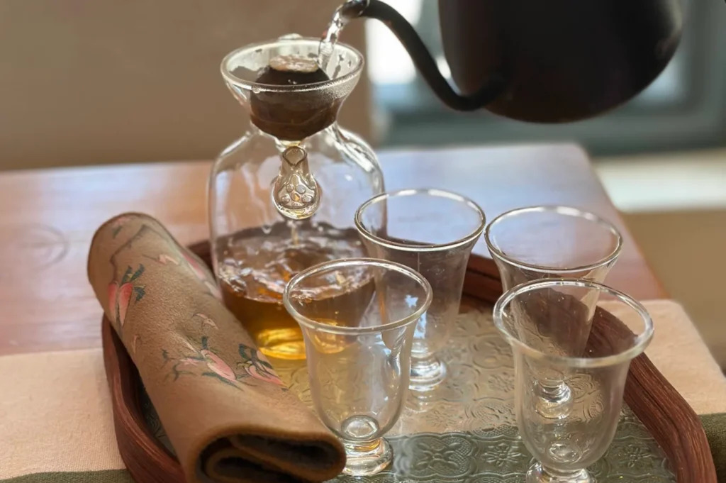 A close-up, elevated shot displays a delicate tea service arranged on a wooden tray, featuring a glass vessel holding golden-amber tea, three matching clear glass cups, and a rolled-up, patterned cloth napkin resting beside the glassware. The scene is captured in soft, natural light, as a dark-colored teapot pours tea into the glass vessel, emphasizing the elegance and quiet sophistication of a modern, minimalist tea ritual.