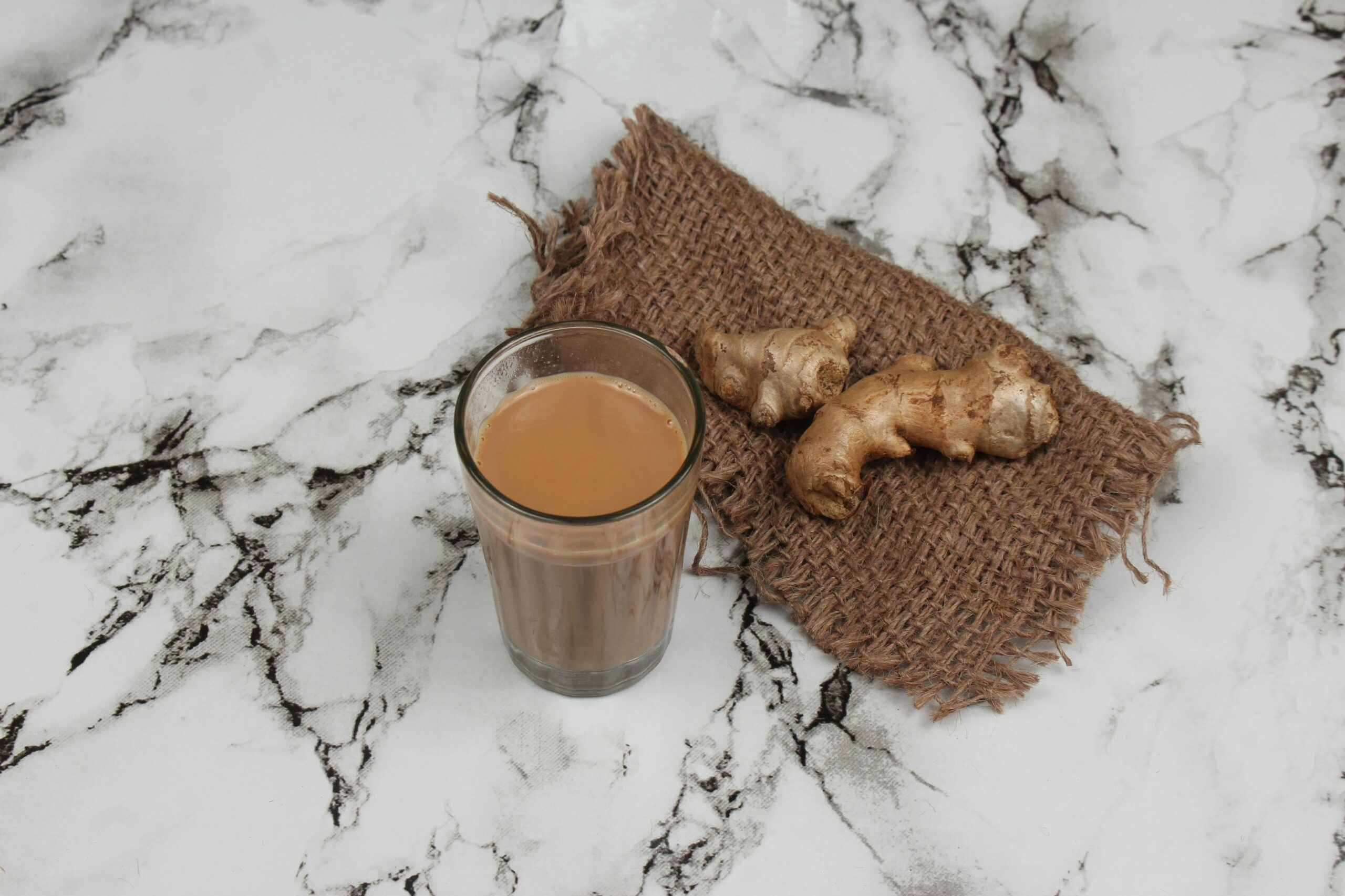 A glass of milky Ginger tea sits on a marble surface next to fresh ginger roots resting on a piece of brown burlap.