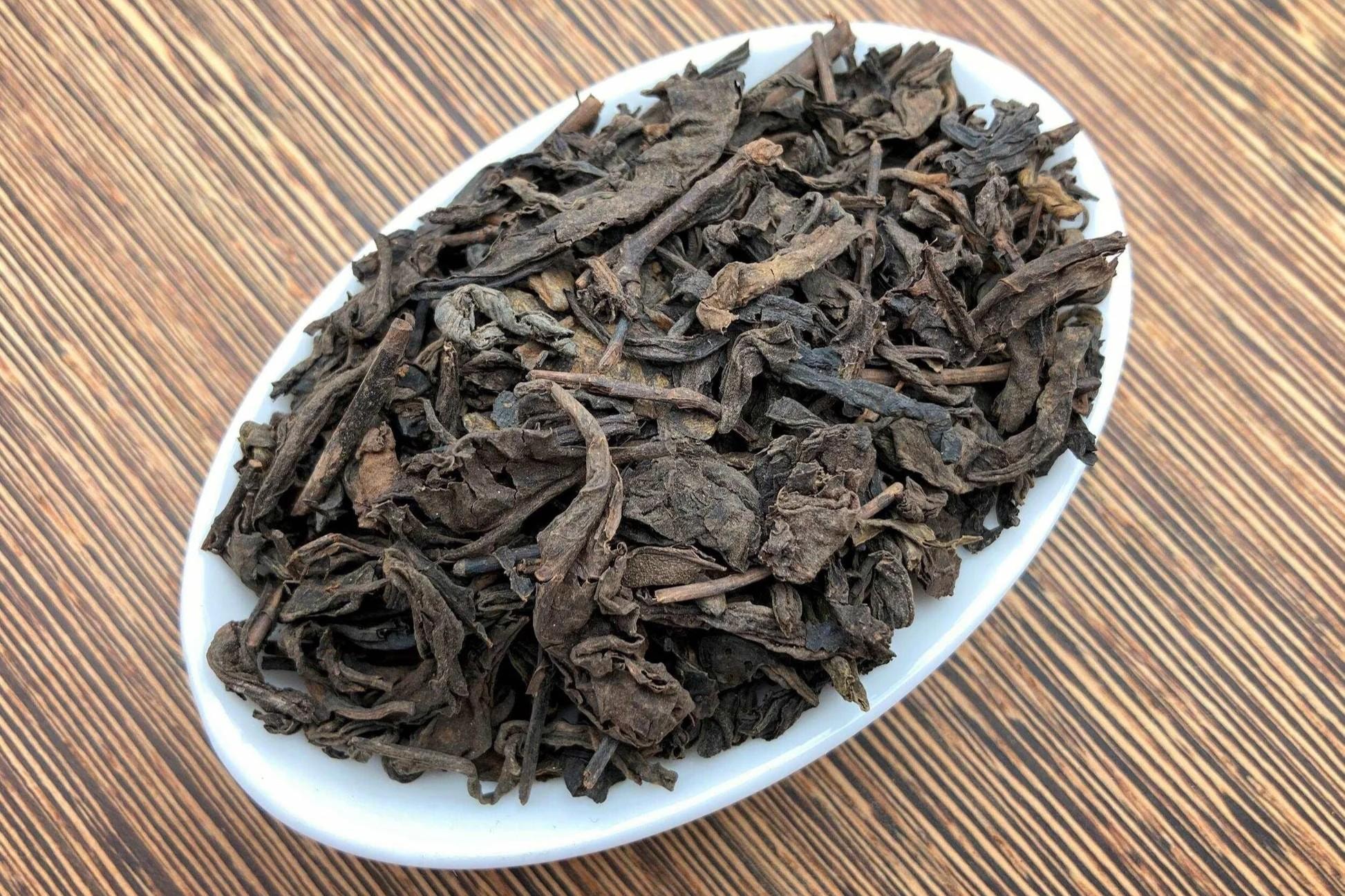 Dried, dark brown Pu-erh tea leaves are piled neatly in a white, oval-shaped ceramic dish. The dish rests on a wooden surface with a distinct, fine-lined grain pattern.