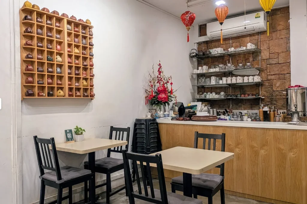 The warm, inviting interior of a teahouse is displayed, characterized by a white wall adorned with a large grid-style wooden display rack filled with small, varied ceramic teapots. The room features simple tables with black chairs, a light-wood service counter backed by a textured stone wall with open shelving for tea sets, and is accented by vibrant red paper lanterns and a floral arrangement, creating a cozy and culturally rich atmosphere.