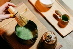 A person with yellow-painted fingernails holds a traditional bamboo whisk over a dark ceramic bowl filled with frothy green matcha tea. The scene is set on a wooden tray and includes other tea ceremony accessories, such as a small lidded jar and a wooden scoop.