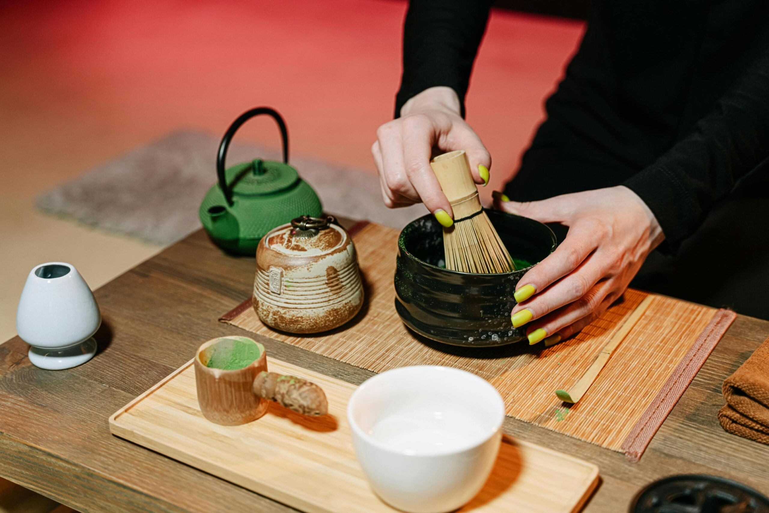  A person with bright yellow nails is shown whisking matcha powder in a black ceramic bowl using a traditional bamboo whisk. The wooden table is neatly arranged with various tea ceremony tools, including a green cast-iron teapot, a small lidded jar, and a white bowl filled with water.