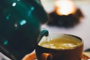A stream of clear tea pours from a teal ceramic teapot into a rustic, earth-toned cup. The close-up shot uses a shallow depth of field to create a warm, cozy atmosphere with soft bokeh in the background.