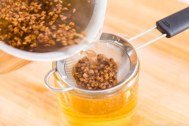 Golden buckwheat tea is being poured from a pot into a glass through a fine-mesh metal strainer. The strainer catches the softened, roasted buckwheat groats while the clear, amber-colored liquid fills the glass below.