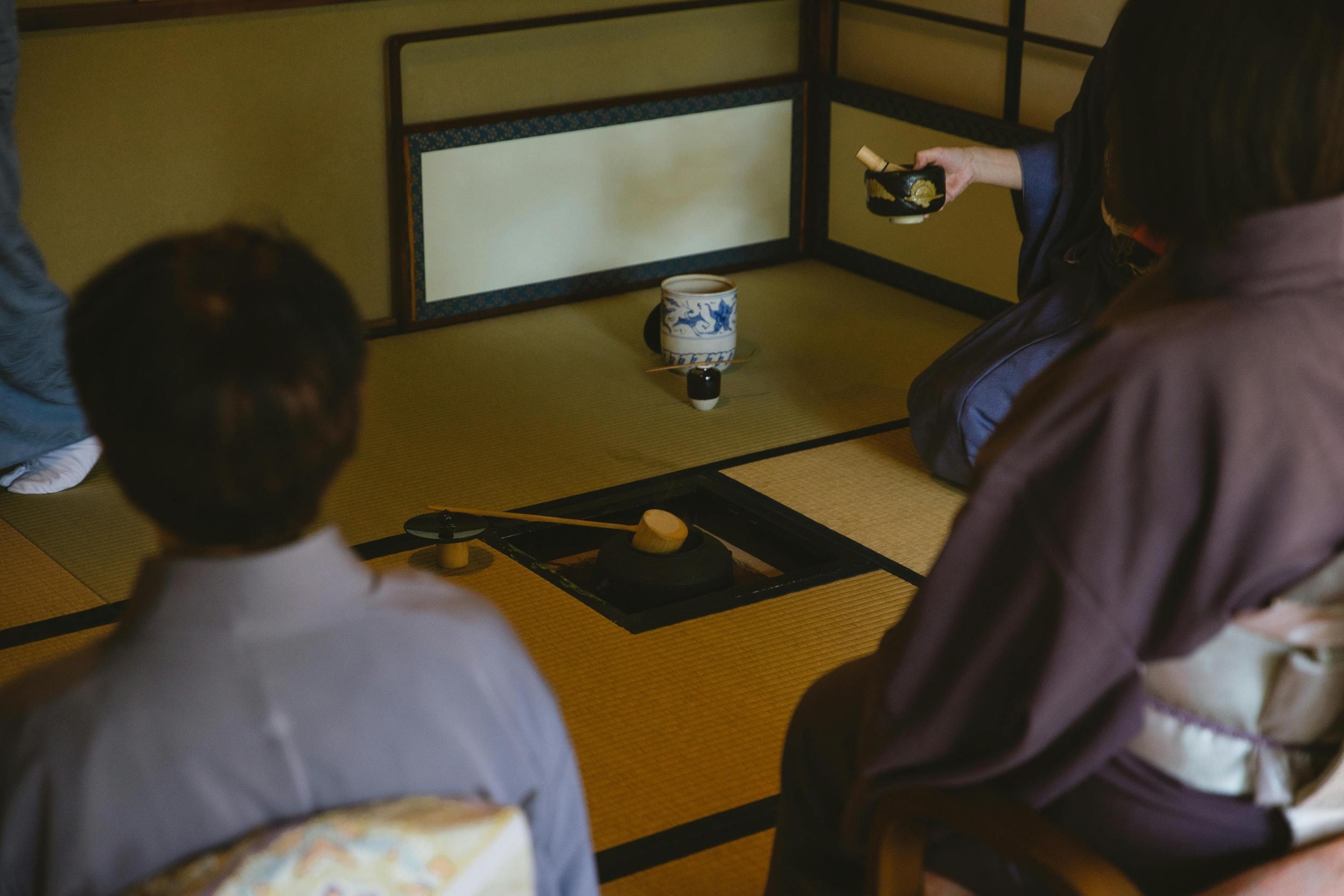 Several individuals dressed in traditional kimonos are gathered on a tatami mat floor for a Japanese tea ceremony. A person in the background holds a tea bowl and whisk, while a sunken hearth with a ladle sits at the center of the scene.
